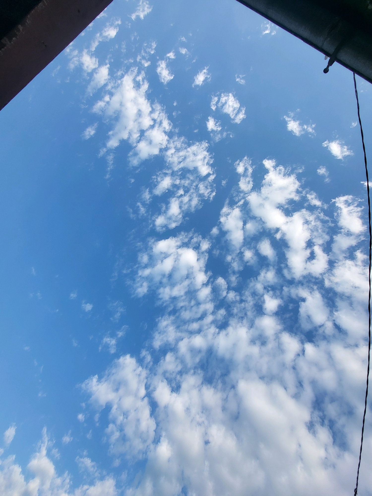The image captures a bright, blue sky with scattered white clouds. A dark, angled structure, possibly part of a building or overhang, is visible in the top left corner. A thin black wire hangs vertically down the right side of the frame. The scene is outdoors and appears to be during the daytime. The location is identified as the city of Jelgava, Latvia. There are no people, animals, or specific activities depicted in the image.