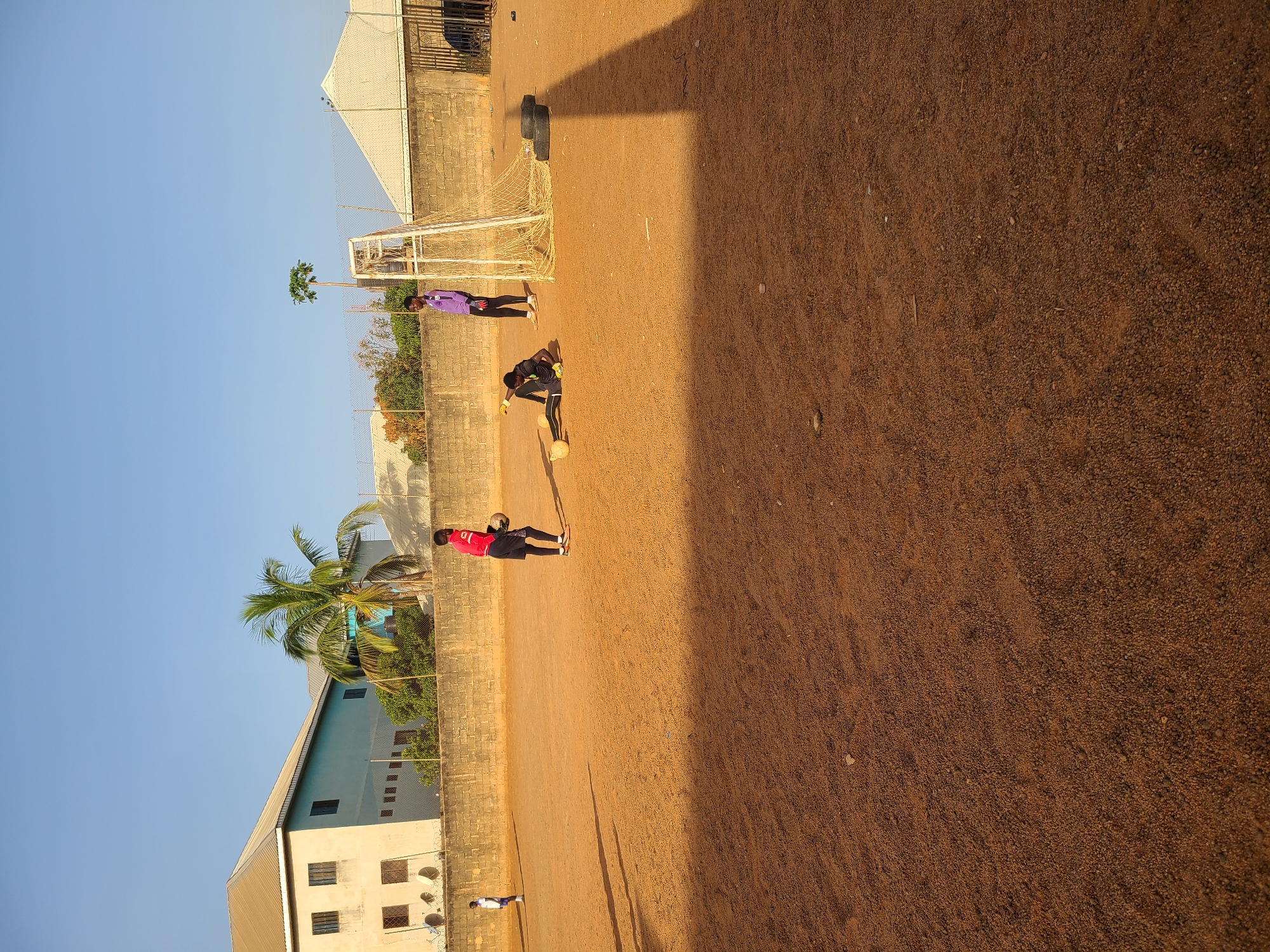 The image shows a soccer field in Jos, Nigeria. The field is made of packed dirt. There are three people visible on the field. One person is wearing a red jersey and is kicking a soccer ball. Another person is wearing a black jersey and is in a goalie position. The third person is wearing a purple shirt. A soccer goal is behind them. A building with a fence is behind the goal. A blue and white building is on the left side of the image with a palm tree in front of it. The sky is blue.