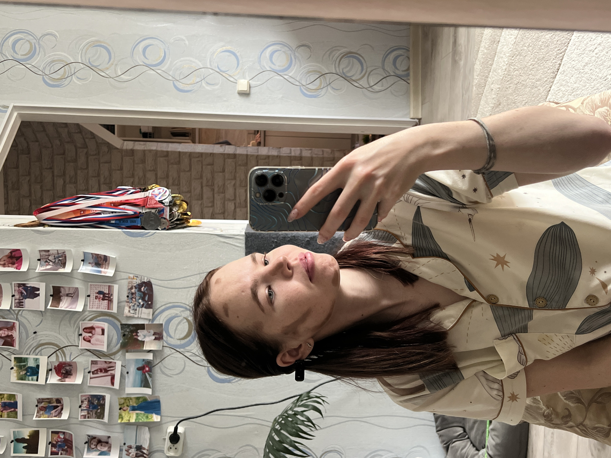 The image is a mirror selfie featuring a young woman taken indoors, likely in a bedroom or personal living space in Chaikovsky, Russia. The photo is rotated 90 degrees clockwise.
The subject is a young woman with shoulder-length dark brown hair, looking directly into the camera of her smartphone. She has several dark smudges or marks on her forehead and left cheek, suggesting recent activity like crafting, painting, or a playful event. She wears a light beige or cream-colored collared shirt with a pattern of dark grey stylized shapes (possibly whales or abstract designs) and small star motifs. The shirt has light brown trim and visible wooden buttons. On her right wrist, she wears a thin silver-colored bracelet. She holds a grey smartphone, likely an iPhone due to its camera array, in her left hand, which has subtly manicured nails.
The background, reflected in the mirror, shows a wall covered in light-colored wallpaper featuring a repeating pattern of wavy lines and circular designs in blue, gold, and grey tones. A collection of small, rectangular printed photos are arranged in a grid on this wall, seemingly personal snapshots. A white electrical outlet with a black cord plugged in is visible near the bottom left of the photo grid. On a shelf or surface, also reflected, to the upper left of the woman's head, is a cluster of medals or awards with colorful ribbons, including one clearly displaying the red, white, and blue colors of the Russian flag, with partially visible text that appears to say "BIG RACE". Other metallic objects and coins are interspersed among the medals. Further back, a grey upholstered furniture item (possibly a couch or chair) and a wall with a textured, brick-like pattern are visible. A white light switch is also on the wall. A green leafy plant is partially visible at the bottom center.
The activity depicted is the casual act of taking a mirror selfie. The woman's expression is neutral, and the indoor setting suggests it could be any time of day.