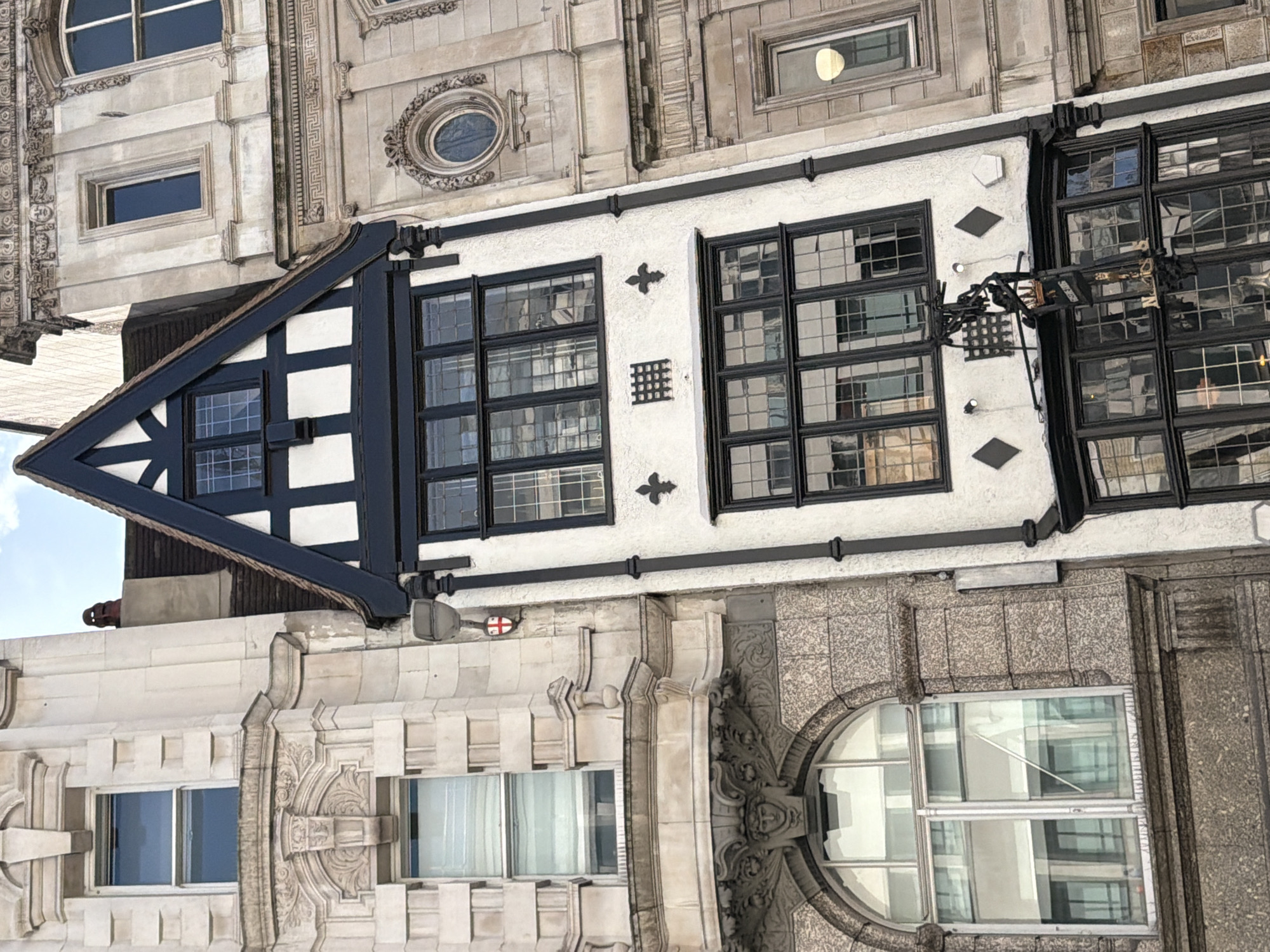The image shows a building in the city of London, United Kingdom. The architecture is a mix of styles, with elements of Tudor Revival and classical design. The building's facade features white stucco with black timber framing, large multi-paned windows, and decorative elements such as crosses and diamond shapes. The lower levels of the building exhibit ornate stonework with rounded windows. The sky is visible behind the building, suggesting a daytime setting. No people are visible.