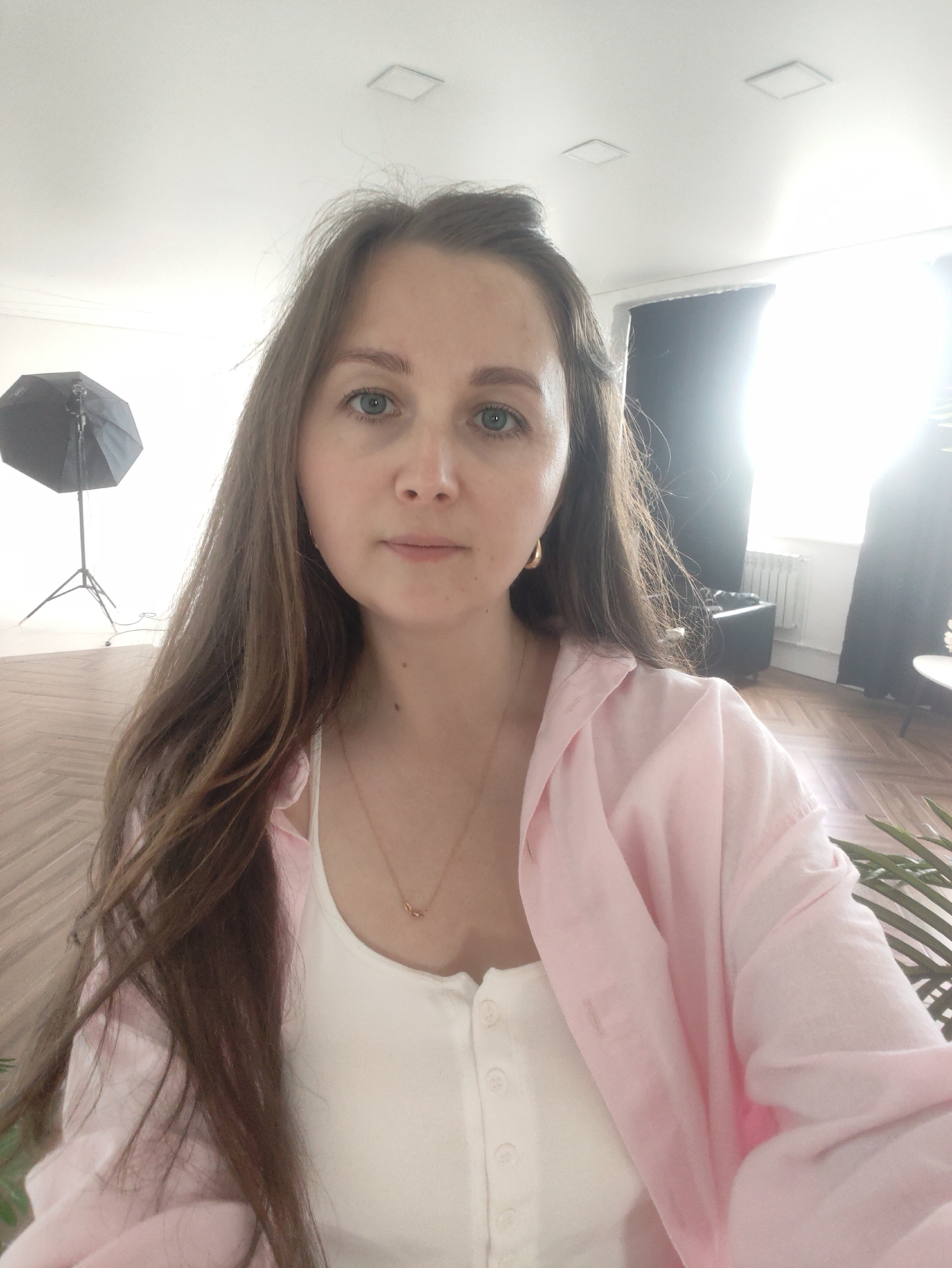 A young woman with long brown hair and blue eyes is taking a selfie in a brightly lit room. She is wearing a white tank top and a pink open shirt. A gold necklace with a small pendant is visible around her neck. In the background, a large umbrella light stands to the left, and dark curtains hang on the right side of the room. The floor is made of wooden planks.