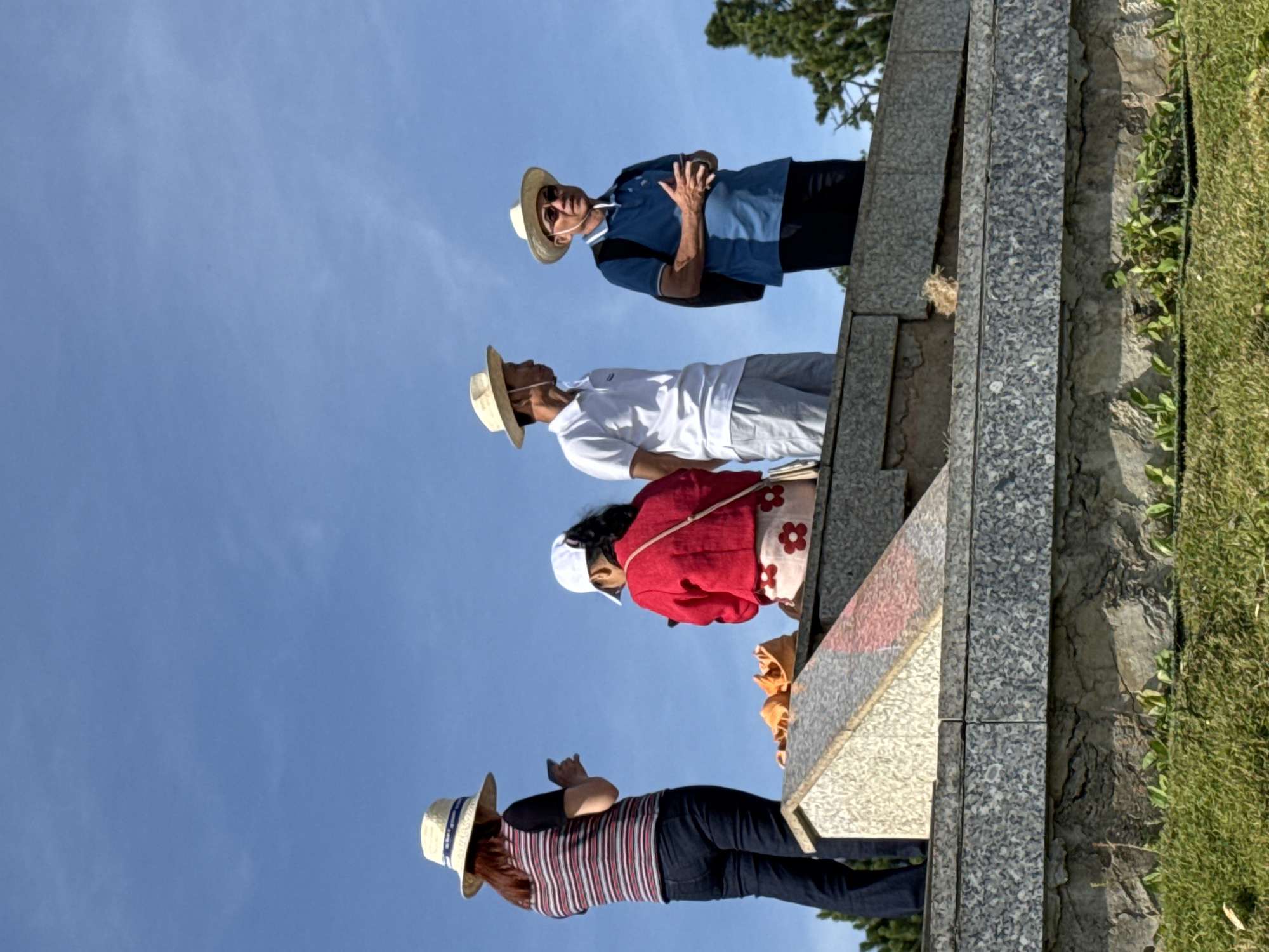 A group of four people, two women and two men, are standing on a tiered stone structure under a clear blue sky in Da Lat, Vietnam. The woman on the far left, wearing a striped shirt and jeans, is looking at her phone. Next to her, a woman in a red jacket and white cap is also looking at her phone. The two men are standing further to the right. The man on the left is wearing a white polo shirt and light-colored shorts, and the man on the right is wearing a blue polo shirt, dark pants, and sunglasses. All four are wearing straw hats. The stone structure appears to be a monument or a memorial, with grassy areas in the foreground and trees visible in the background.