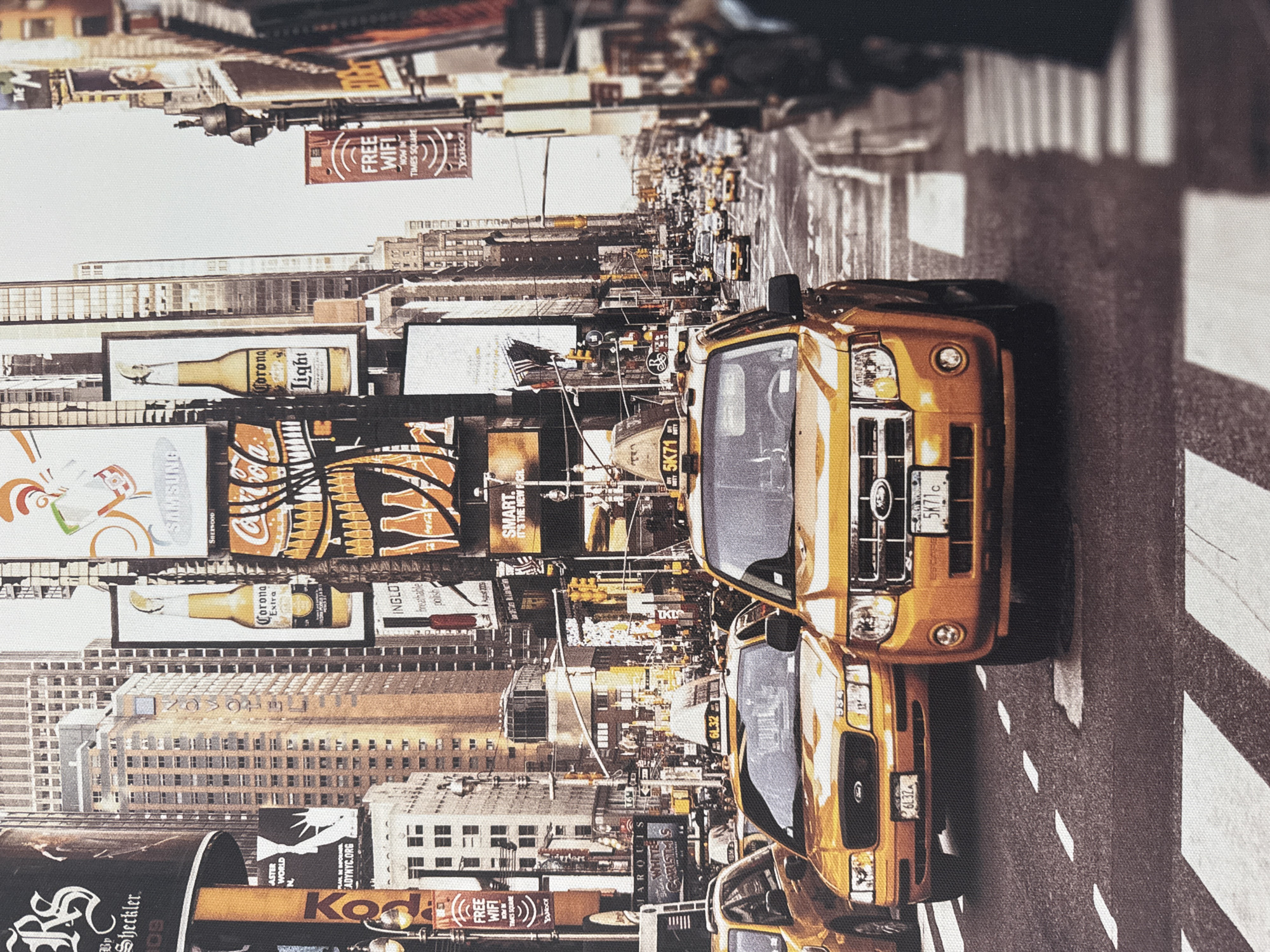 This image depicts a busy city street scene, likely in Times Square, New York City, characterized by towering buildings adorned with numerous large billboards and advertisements. Yellow taxis are prominently featured, with several in the foreground and more visible in the background, suggesting active traffic. The architecture and the style of the advertisements, including brands like Corona and Coca-Cola, are indicative of a major urban center. The environment appears to be a bright day, possibly late afternoon, given the lighting. The scene is filled with vehicles and the general bustle of city life, with pedestrians blurred in the background. The location is *not* Анкудиновка, Russia.