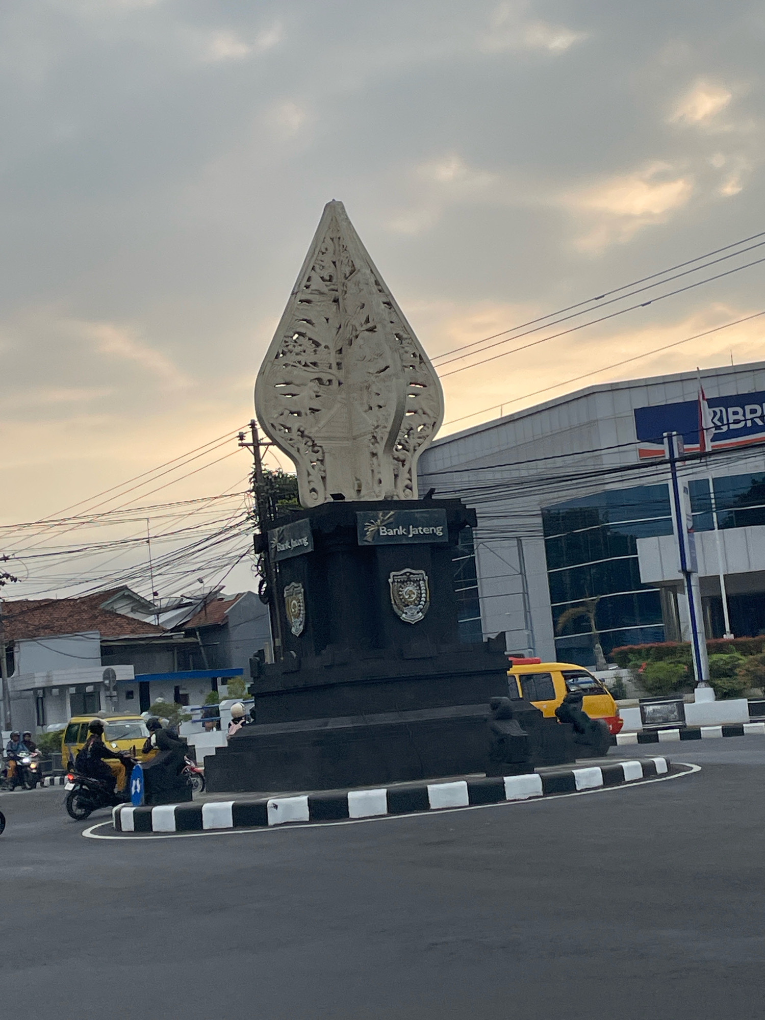The image depicts a street scene in Purworejo, Indonesia, likely at a roundabout. Dominating the center is a large, white, intricately carved sculpture atop a black, tiered base. The sculpture's design is abstract and pointed, resembling a stylized flame or traditional Javanese headdress. On the black base, text reading "Bank Jateng" is visible on two sides, alongside emblems.
Surrounding the roundabout, several vehicles are present. A yellow minibus with a red light on top is visible. Several motorcycles are also in motion, with riders dressed in various casual clothing, some wearing helmets. One motorcycle has a rider wearing black and a passenger also in dark clothing. Another motorcycle has a rider in a yellow shirt.
In the background, a modern building with glass windows and a blue sign that says "BRI" is prominent. Utility poles and a dense network of electrical wires crisscross the sky. Adjacent to the BRI building, a smaller building with a red-tiled roof is visible. The sky is overcast with diffused light. The road surface is asphalt, and the roundabout is bordered by black and white striped curbs.