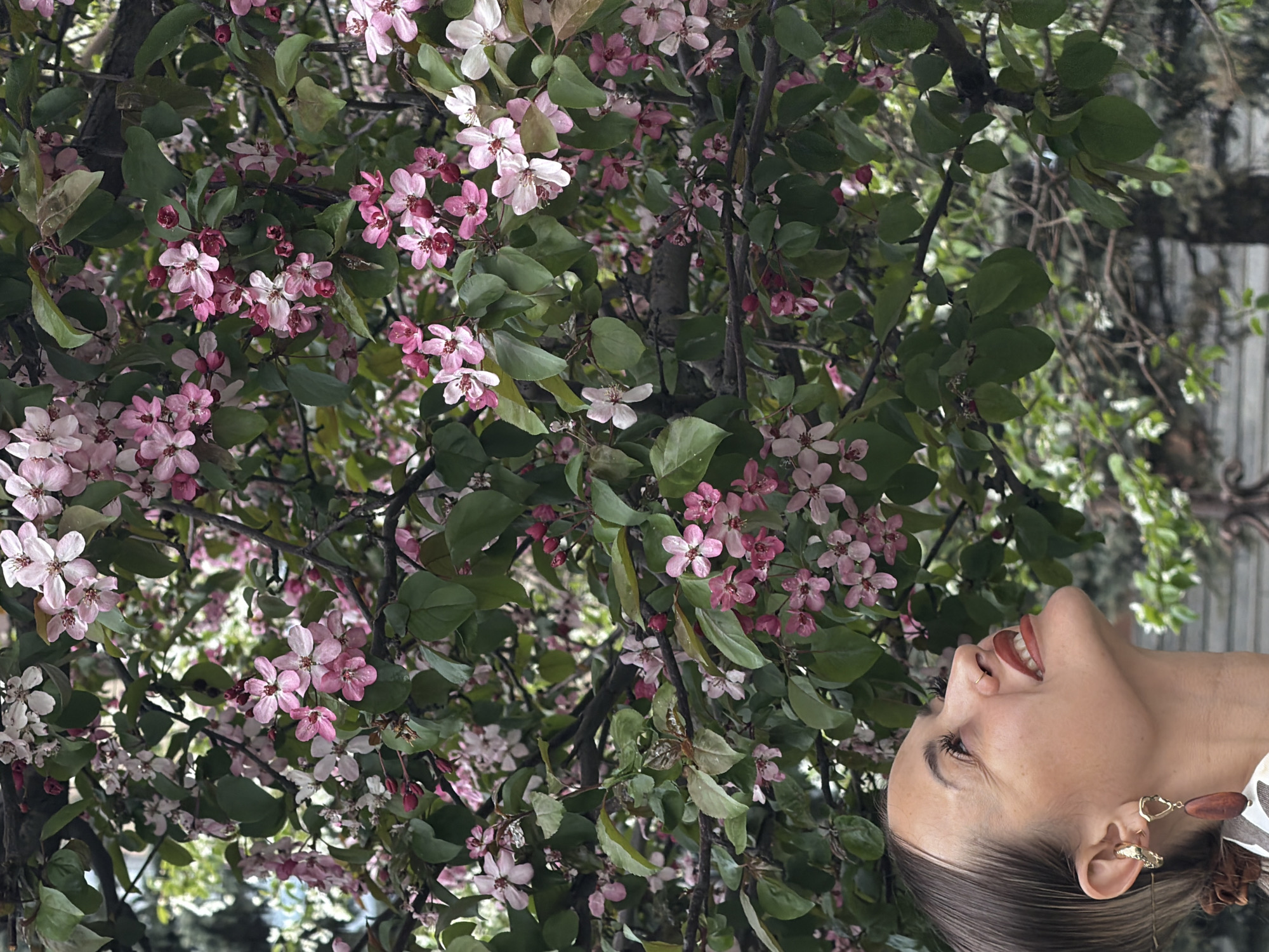 A medium shot shows a woman looking upward, smiling at a profusion of pink and white blossoms. She is right of center in the frame. She has dark hair pulled back, and her right ear is visible with a gold earring. The backdrop is a dense canopy of flowers, likely in a tree. The city of Krasnoyarsk, Russia, is not evident in the image.