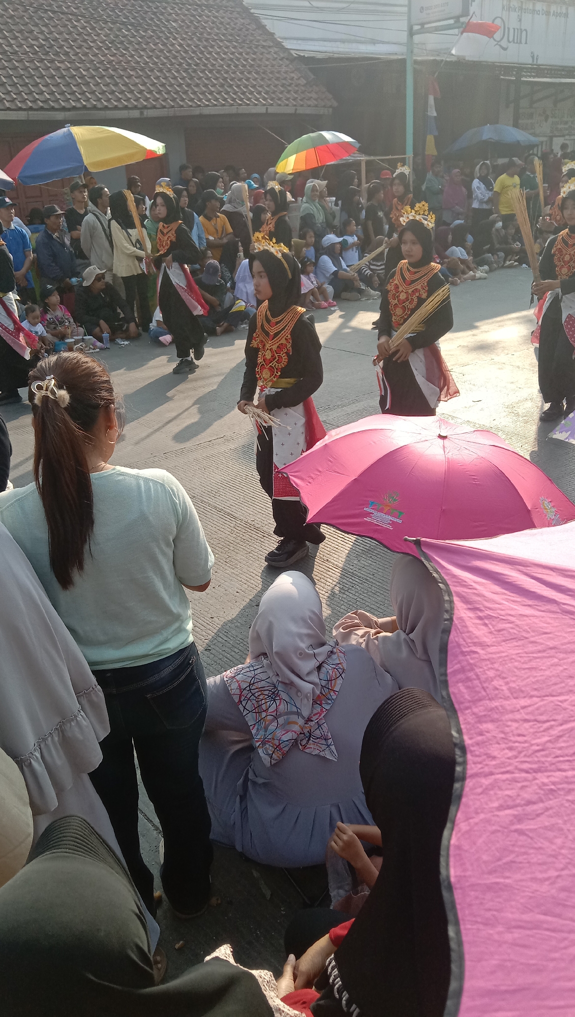In Jatisawit, Indonesia, a street parade is underway with participants in traditional attire and onlookers. The event is taking place outdoors on a paved surface, likely a street, with buildings and trees visible in the background. The sun is shining, casting long shadows.
The central figures of the parade are young women dressed in elaborate, dark costumes adorned with gold embroidery on the chest and headpieces. They are holding bundles of dry grass or straw. One woman in the foreground, dressed in similar ceremonial attire, is looking towards the camera while holding the straw. To her right, another participant is walking away from the camera.
A diverse crowd of spectators is gathered on both sides of the parade route. Many are seated or standing, observing the procession. Umbrellas of various colors, including red, yellow, blue, and green, are being used to provide shade. A large pink umbrella is prominently featured in the foreground, partially obscuring the view of some spectators.
Several individuals in the foreground are seated, facing away from the camera, and appear to be watching the parade. One woman in a light blue shirt and jeans is looking towards the parade participants. The overall atmosphere suggests a cultural celebration or festival.