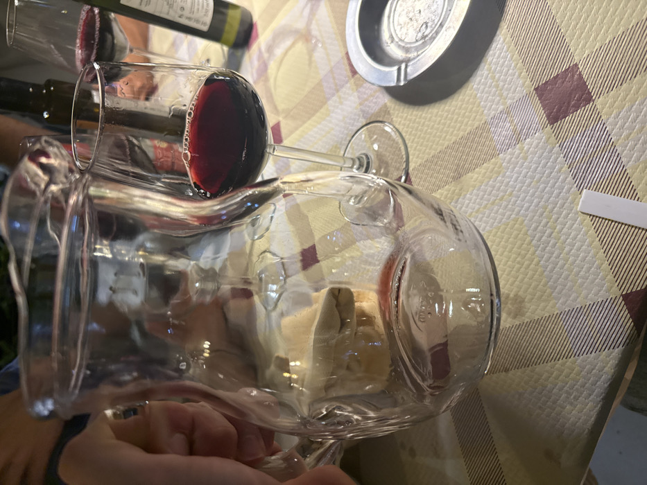 An indoor scene in a dining establishment in Lecce, Italy, displays a portion of a checkered tablecloth featuring light yellow, beige, and maroon patterns. In the foreground, a clear glass pitcher is held by a left hand positioned on the left side of the frame. The pitcher, appearing mostly empty with a small amount of liquid at the bottom, reflects various blurred objects from the background, including other glass vessels and indistinct forms. To the immediate right of the pitcher, a wine glass filled with red wine stands on the tablecloth, exhibiting small bubbles at the liquid's surface. Further to the right and slightly behind, another wine glass is partially visible, also containing red wine. A dark bottle and a green wine bottle (with "750 ml" visible on its label) are present in the background. On the right side of the table, a circular metallic ashtray containing dark residue and possibly a cigarette butt is visible. A small white rectangular slip of paper rests near the bottom center of the frame. Through the pitcher, a light-colored, possibly edible, object like bread can be discerned on the table. The overall scene depicts a casual dining or social setting.