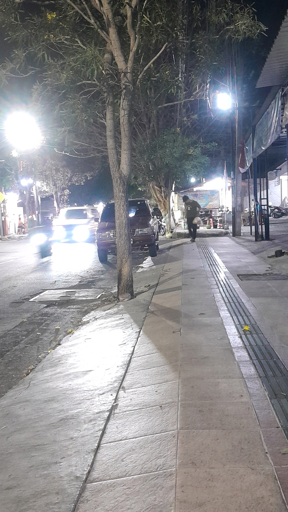The image depicts a street scene at night in Madiun, Indonesia. A sidewalk with tactile paving runs along the right side of the frame, parallel to the street. A single person is walking away from the viewer on the sidewalk, carrying an object. To the left of the sidewalk, a street is visible with two cars, one of which has its headlights on, illuminating the road. A tree with sparse foliage stands between the sidewalk and the street, casting shadows. In the background, there are buildings, streetlights, and more trees. The overall atmosphere is that of a dimly lit urban environment at night.