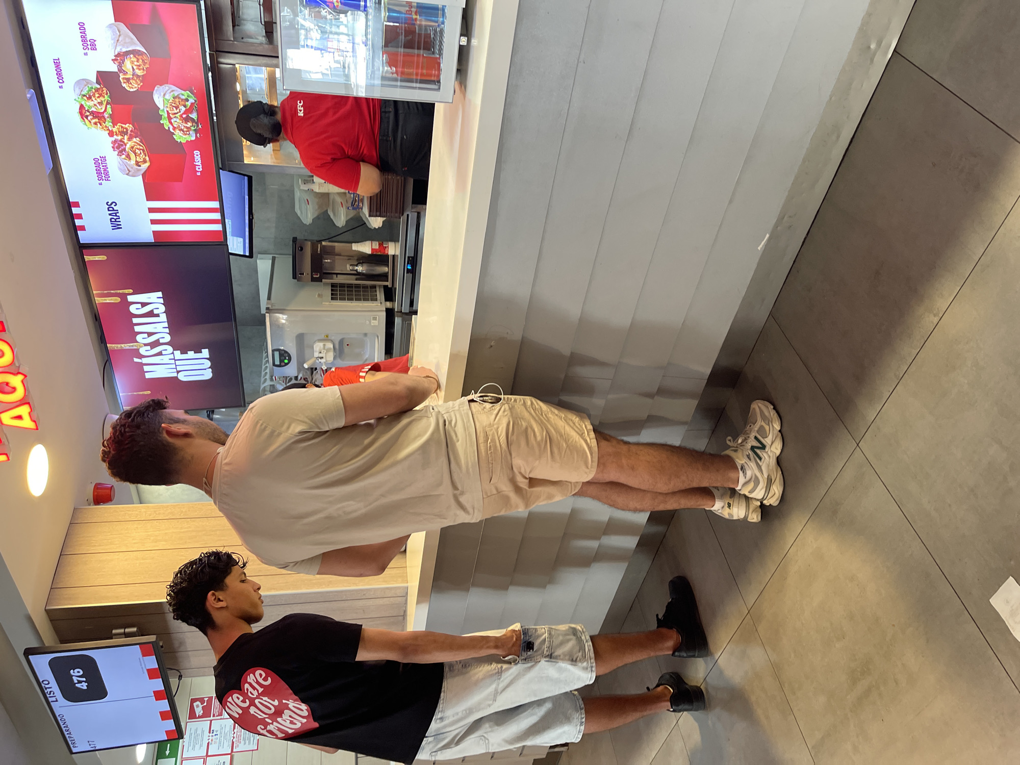 The image depicts the interior of a KFC restaurant, likely in Barcelona, Spain, given the Spanish text on the screens. Two young men are standing at the counter, facing away from the camera. The man on the left wears a black t-shirt with a red heart graphic that says "we are not friends" and light-colored cargo shorts. The man on the right wears a light-colored t-shirt and tan shorts. A KFC employee in a red uniform is visible behind the counter, interacting with a customer not fully in frame. Above the counter, large screens display menu items, including wraps, with Spanish text. To the left, a digital display shows "PREPARANDO LISTO" and the number "476". The environment appears clean and well-lit, with tiled flooring and white paneled walls.