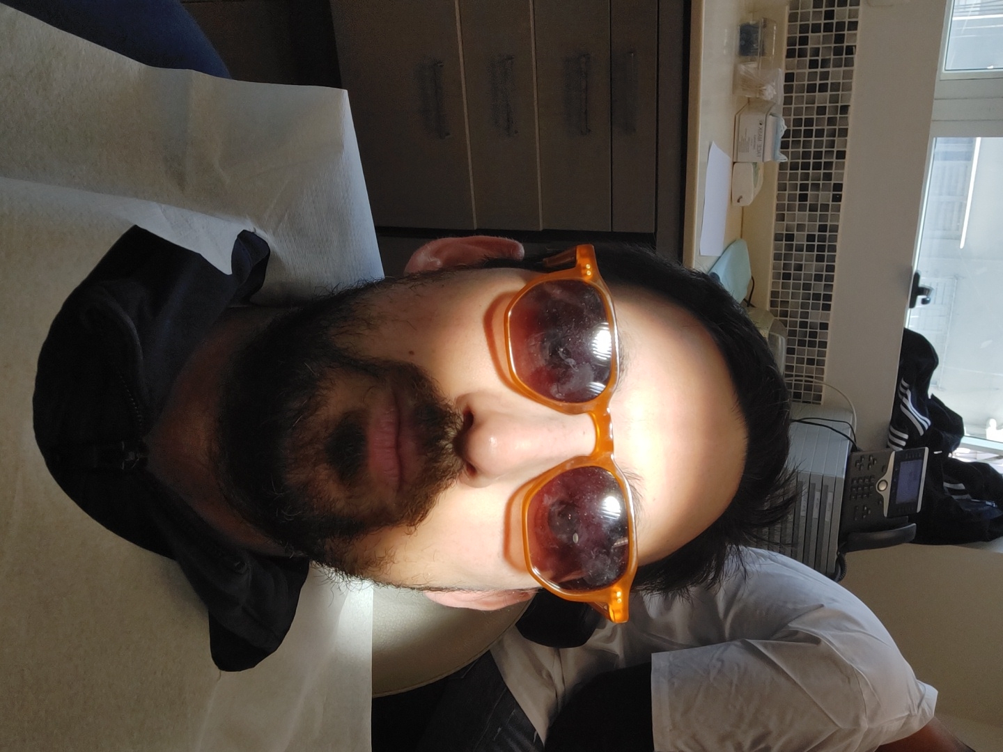 A man with a beard is lying on a medical examination table in an office setting in Tel Aviv, Israel. He is wearing orange sunglasses. The office has a window showing buildings outside. There is medical equipment in the background. A black jacket lies next to him.
