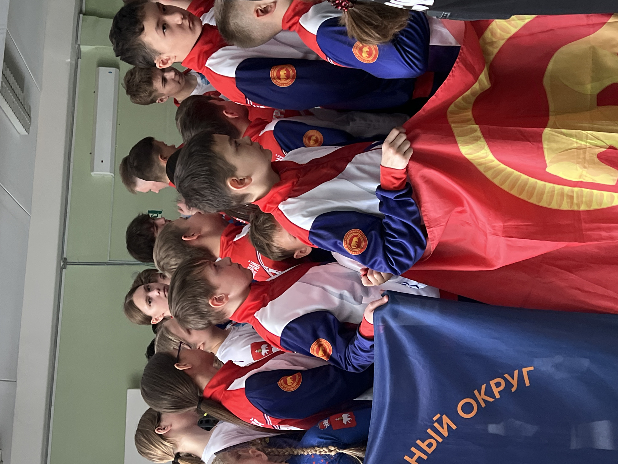 A group of children are standing together, holding a red flag with a yellow emblem and a dark blue flag with orange text. The children are wearing matching tracksuits with a white center, red sleeves and shoulders, and dark blue arms. A patch with a bear design is visible on the sleeves of the tracksuits. The scene appears to be indoors, potentially in Berezniki, Russia.