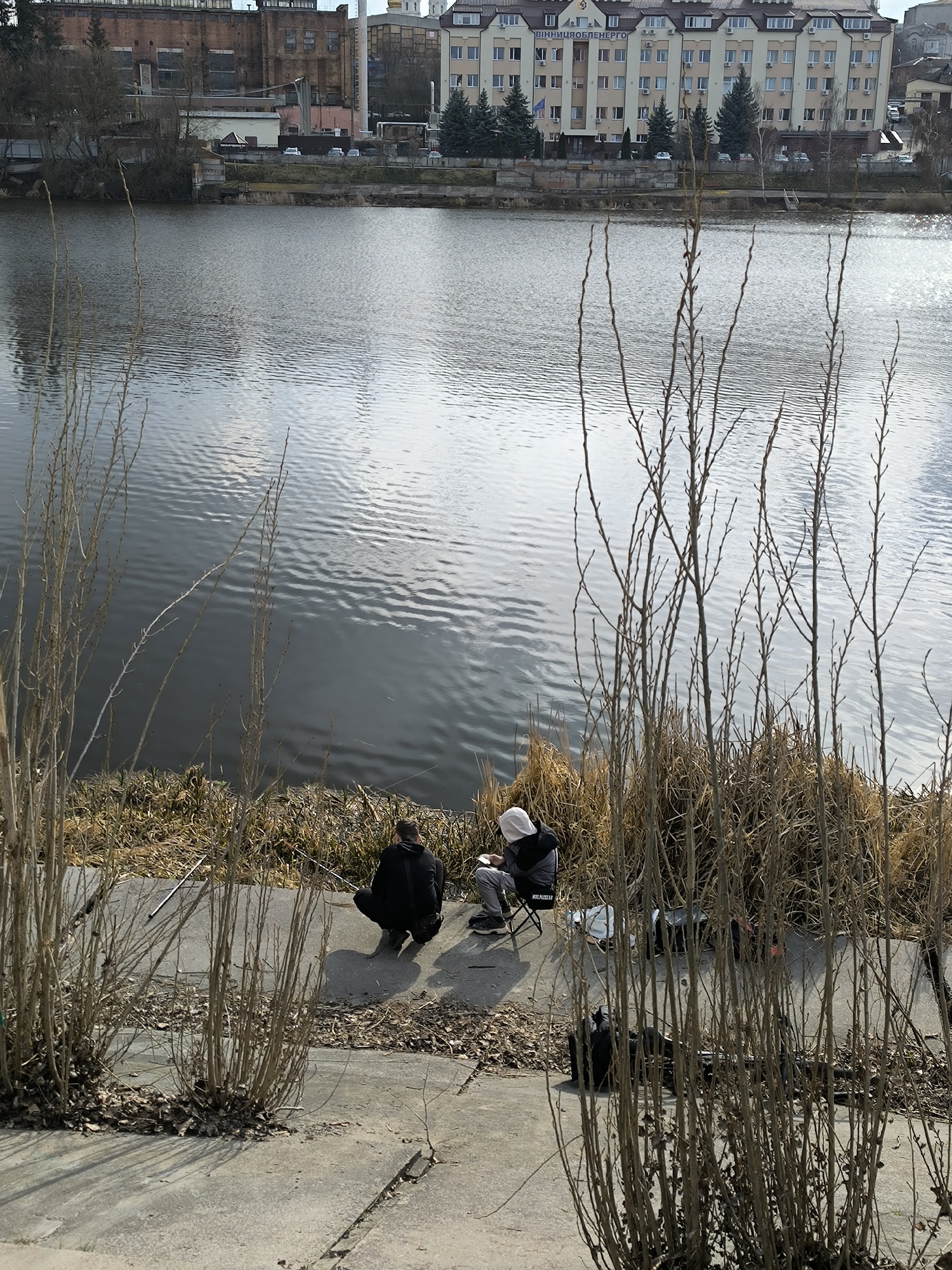 The image depicts two individuals fishing by the Southern Bug river in Vinnytsia, Ukraine. The scene includes the river with reflections, trees on the opposite bank, and buildings in the background. In the foreground, there are sparse bushes and vegetation, obscuring a paved area where the two people are situated. One person is squatting while the other is sitting on a folding chair. Fishing rods are visible. Personal items such as bags are nearby. The weather appears to be overcast.