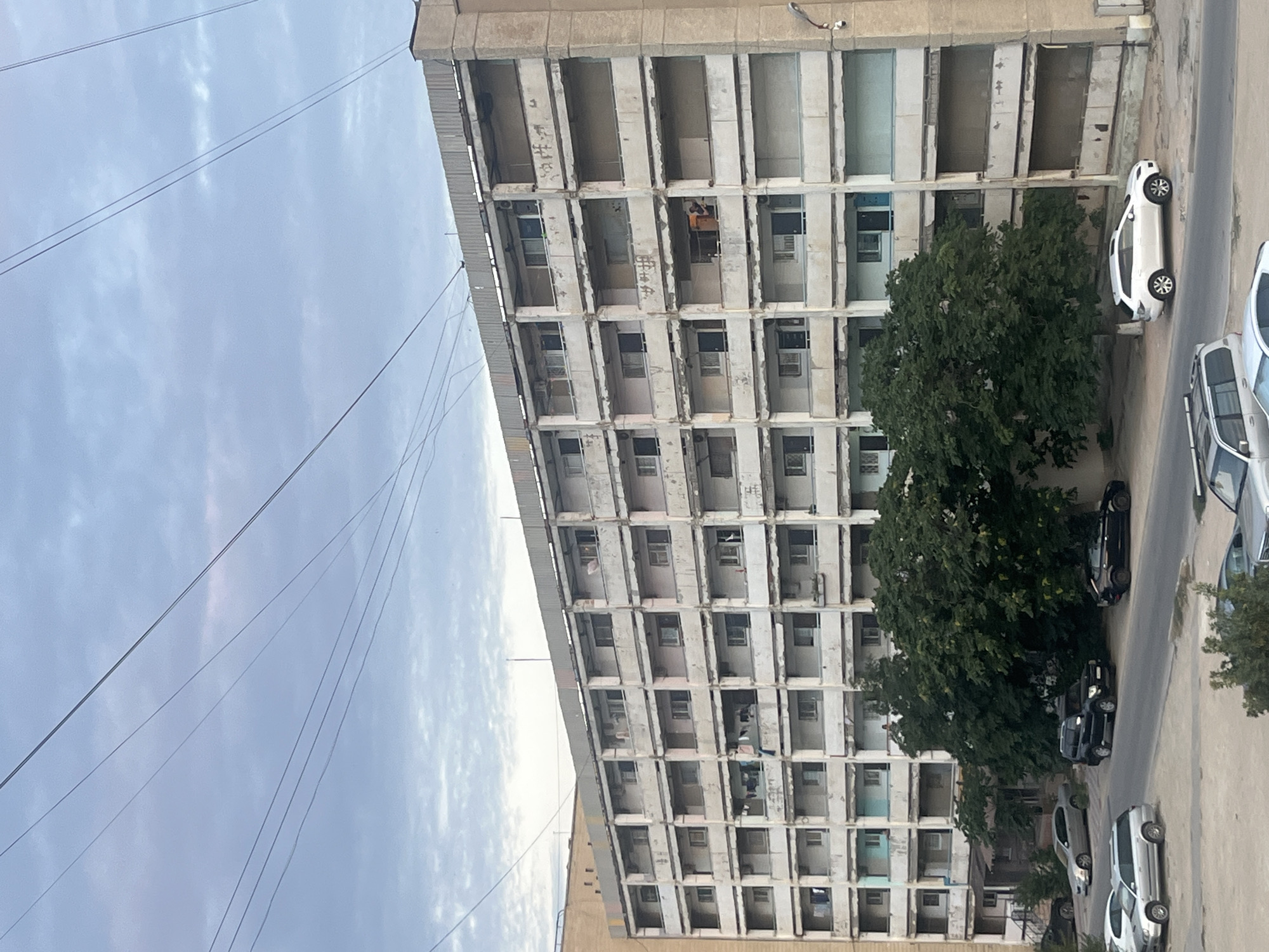 The image depicts a multi-story apartment building in the city of Aktau, Kazakhstan. The building appears to be constructed of concrete and has a weathered facade with many balconies. Laundry is hanging on some of the balconies, and a few people are visible in open doorways or on the balconies. In front of the building, several cars are parked on a street and a dirt area, including a silver sedan, a black SUV, a black sedan, a white SUV, and parts of other vehicles. Large green trees partially obscure the lower floors of the building. The sky above is partly cloudy.
