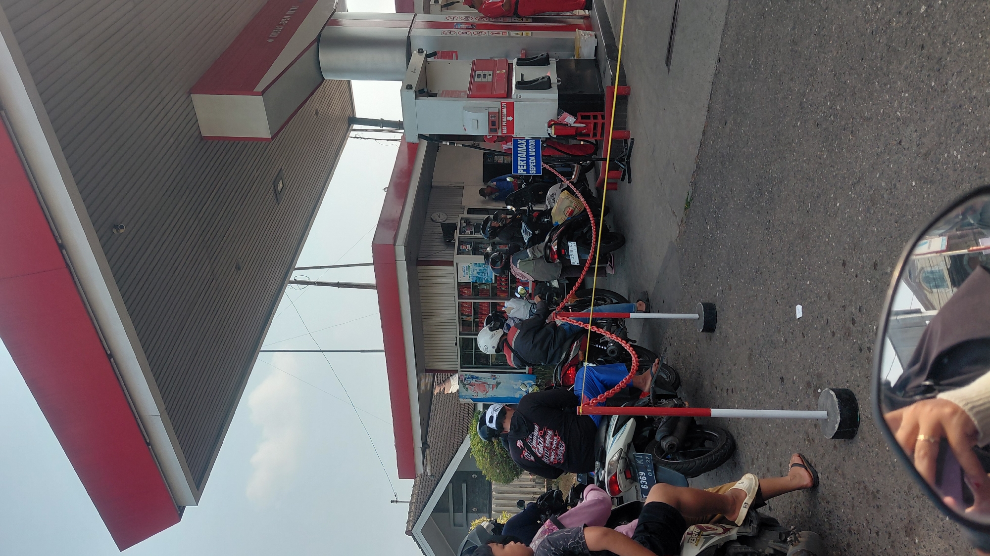 This is a street-level, eye-level, vertical photograph taken at a Pertamina gas station in Rejasari, Indonesia. The scene depicts several individuals on motorcycles waiting in line to refuel. The gas station canopy, painted red and white, extends over the pumps. A white and red gas pump is visible on the right, with a sign that reads "PERTAMAX SEPEDA MOTOR". There are two red and white bollards with chains in the foreground, indicating a queueing area. Some people are wearing helmets, and one person in the foreground appears to be wearing a white and black hooded sweatshirt. The ground is asphalt. In the reflection of a motorcycle's rearview mirror at the bottom of the frame, a person's hand with a ring is visible, along with buildings in the background. The sky is partly cloudy.