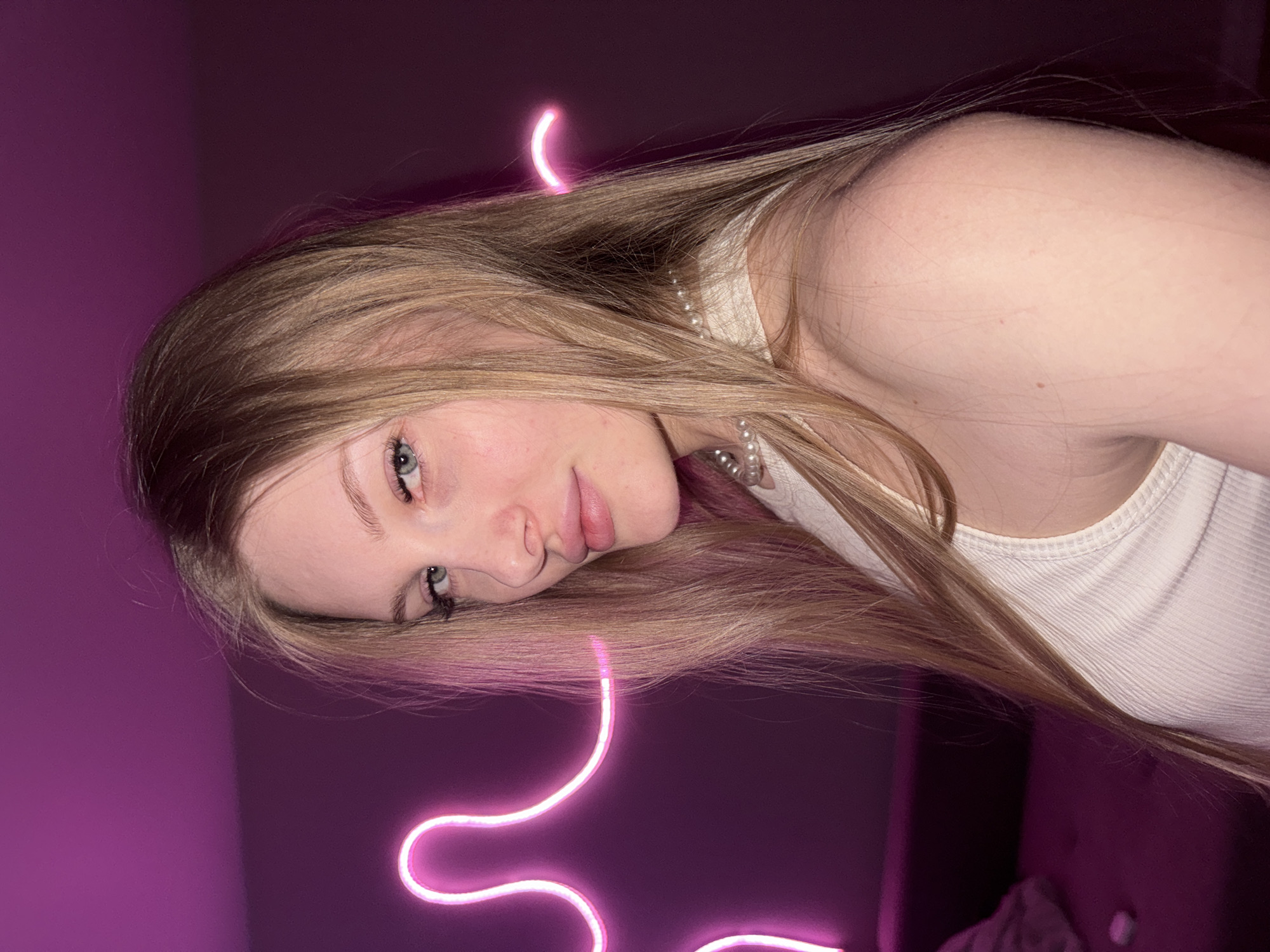 A young adult female with long, straight blonde hair and light blue eyes is depicted from the chest up, looking over her right shoulder directly at the viewer. She wears a white ribbed tank top and a multi-strand pearl necklace. A slight reddish mark is visible on her skin above her right eye. The background features a dark purple wall, brightly illuminated by a pink neon light fixture shaped as a stylized human figure or abstract wavy lines. A portion of a dark sofa or bench is visible in the lower-left corner of the frame, alongside a small, light-colored object on its surface. The overall lighting is dim, primarily emanating from the neon fixture.
