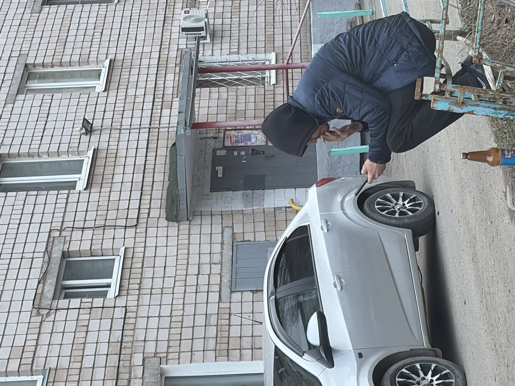 A photograph shows a man squatting near a parked, silver hatchback car next to a brick apartment building. The man wears a dark hooded jacket, dark pants, and dark footwear. He faces towards the car's rear tire, extending his hand towards the tire. A glass bottle lies on the ground near the car. The apartment building behind the man features multiple windows, an air conditioning unit, and a dark doorway. The setting appears to be an urban street in Izhevsk, Russia.