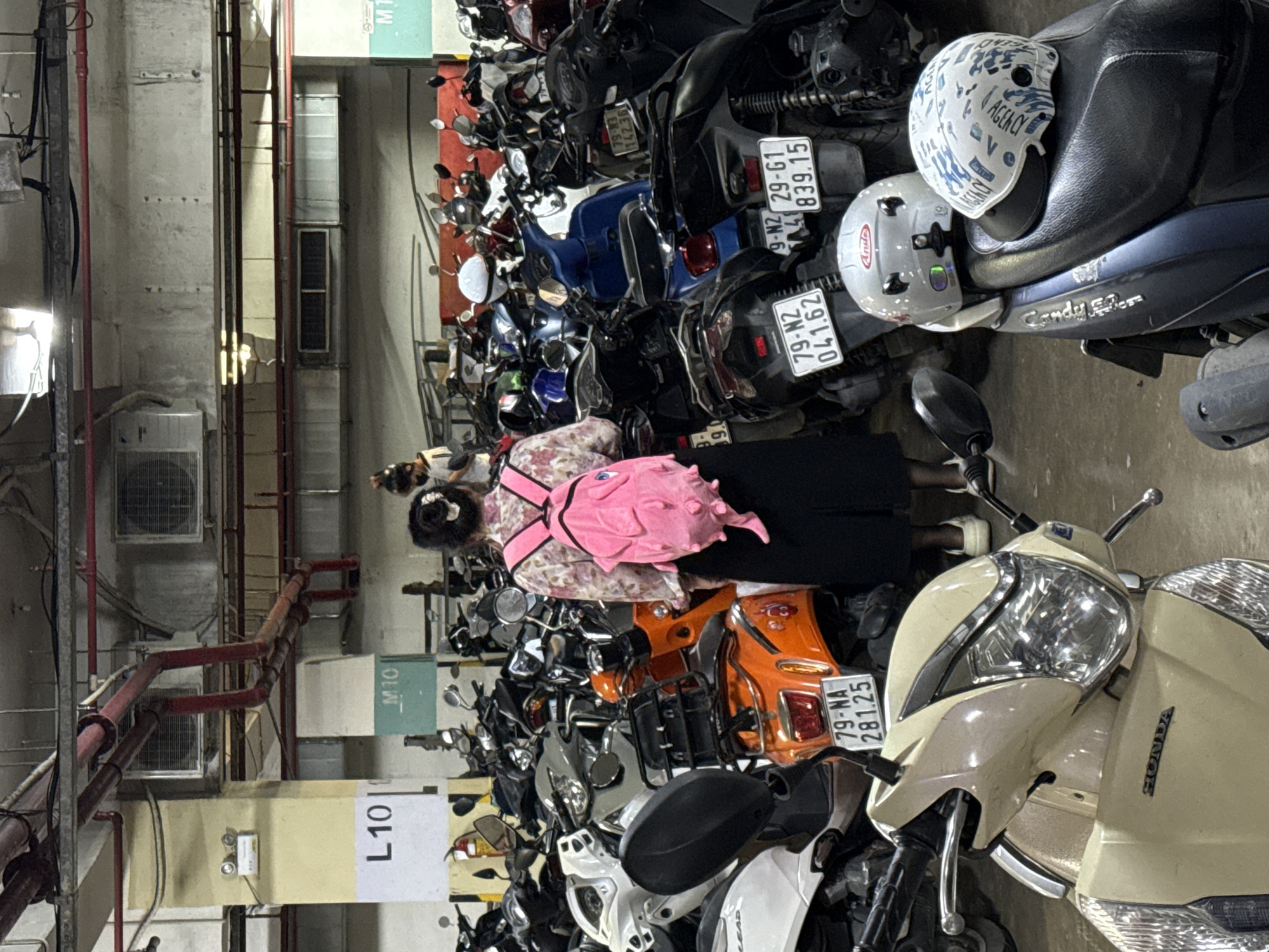 This is a vertical, full shot of a parking garage in Nha Trang Ward, Vietnam. The scene is filled with parked motorcycles and scooters. In the foreground, a woman wearing a floral dress and a pink backpack stands facing away from the camera, appearing to be walking. She is surrounded by numerous parked vehicles. Behind her, more motorcycles and scooters are packed tightly together. Two helmets are visible on the seat of a blue scooter in the middle ground, one white with a blue pattern and the other white with "Ande" branding.
The lighting suggests it is daytime, but the environment appears to be indoors, likely a covered parking area. Several signs are visible: "L101," "M10," and "M1." License plates on some of the motorcycles display text such as "79-NA 281.25," "79-N2 041.62," and "9-N2 29-G1 839.15." The overall impression is a busy, functional space for vehicle storage. No specific event is clearly depicted, but the presence of the woman and the neatly parked vehicles suggests a normal day in a populated area.