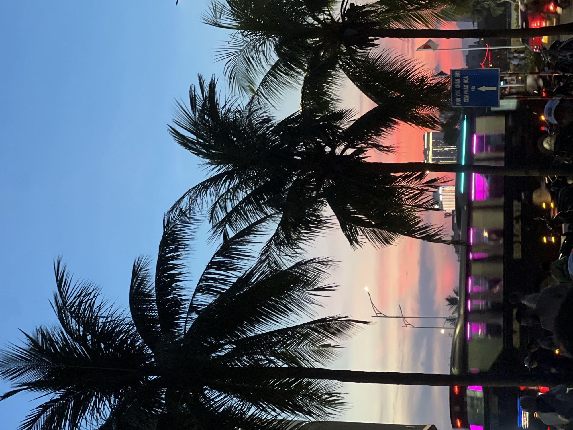 The image captures a vibrant sunset scene in an urban environment, likely a coastal city given the presence of palm trees. The sky transitions from a deep blue at the top to a fiery orange and pink near the horizon, with silhouetted palm trees in the foreground framing the view. A bus with illuminated purple lights moves across the frame, suggesting a bustling street scene. In the background, buildings and streetlights are visible, indicating a developed area. Several people are gathered on what appears to be a street or sidewalk, some on motorcycles. A blue sign with Vietnamese text and an upward arrow is prominently displayed, providing directional information.