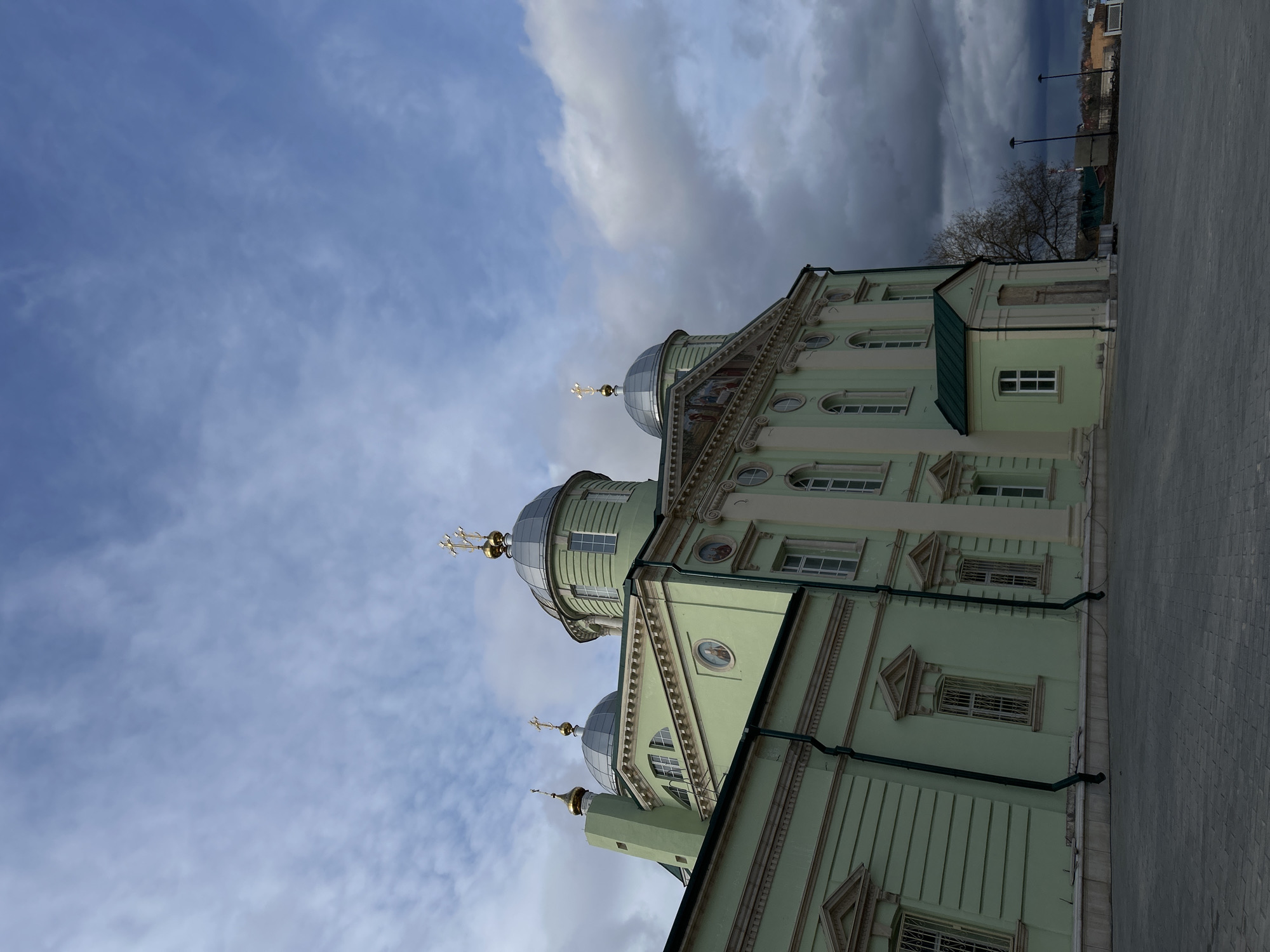 The image shows the Cathedral of the Assumption in the city of Klin, Russia. The light green building has silver domes with golden crosses on top. The sky is blue with white and grey clouds. The building is located next to a brick paved square.