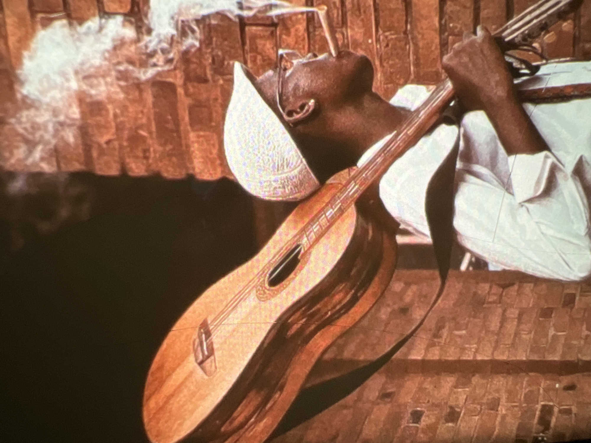 This image captures a close-up of a Black man, possibly a musician, with dark skin and short hair, wearing glasses and a light-colored cap. He has a cigar in his mouth and is looking to the right, with smoke rising around him. He is carrying a guitar over his right shoulder. The man is wearing a white shirt, and a strap from the guitar is visible. He appears to be standing against a brick wall. The overall lighting suggests an outdoor setting, potentially during the day, with the brick wall indicating an urban environment. The image's composition, with the guitar prominently featured and the man's attire, suggests a musical context, possibly a performance or travel. Without specific landmarks or context, it is not possible to definitively place this scene in Lagos, Nigeria.