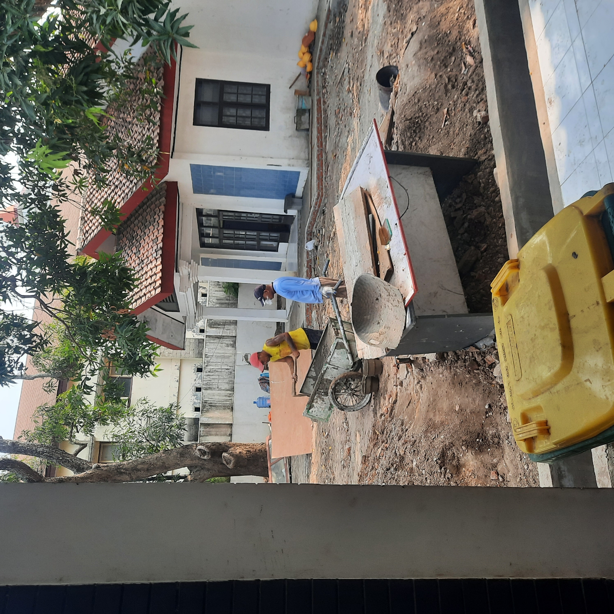 Two men are working in a construction site in front of a white building with a red-tiled roof. One man, wearing a yellow tank top and a red baseball cap, is using a shovel to load material into a wheelbarrow. The other man, in a blue long-sleeved shirt and a blue baseball cap, stands and observes. The ground is covered in dirt and debris, with scattered construction materials like wood planks and bricks. A large tree with dense foliage is visible on the left side of the image, casting shadows. In the foreground, a yellow trash bin is partially visible. The scene suggests ongoing renovation or construction work in an outdoor setting. This location is identified as Lemahabang, Indonesia.