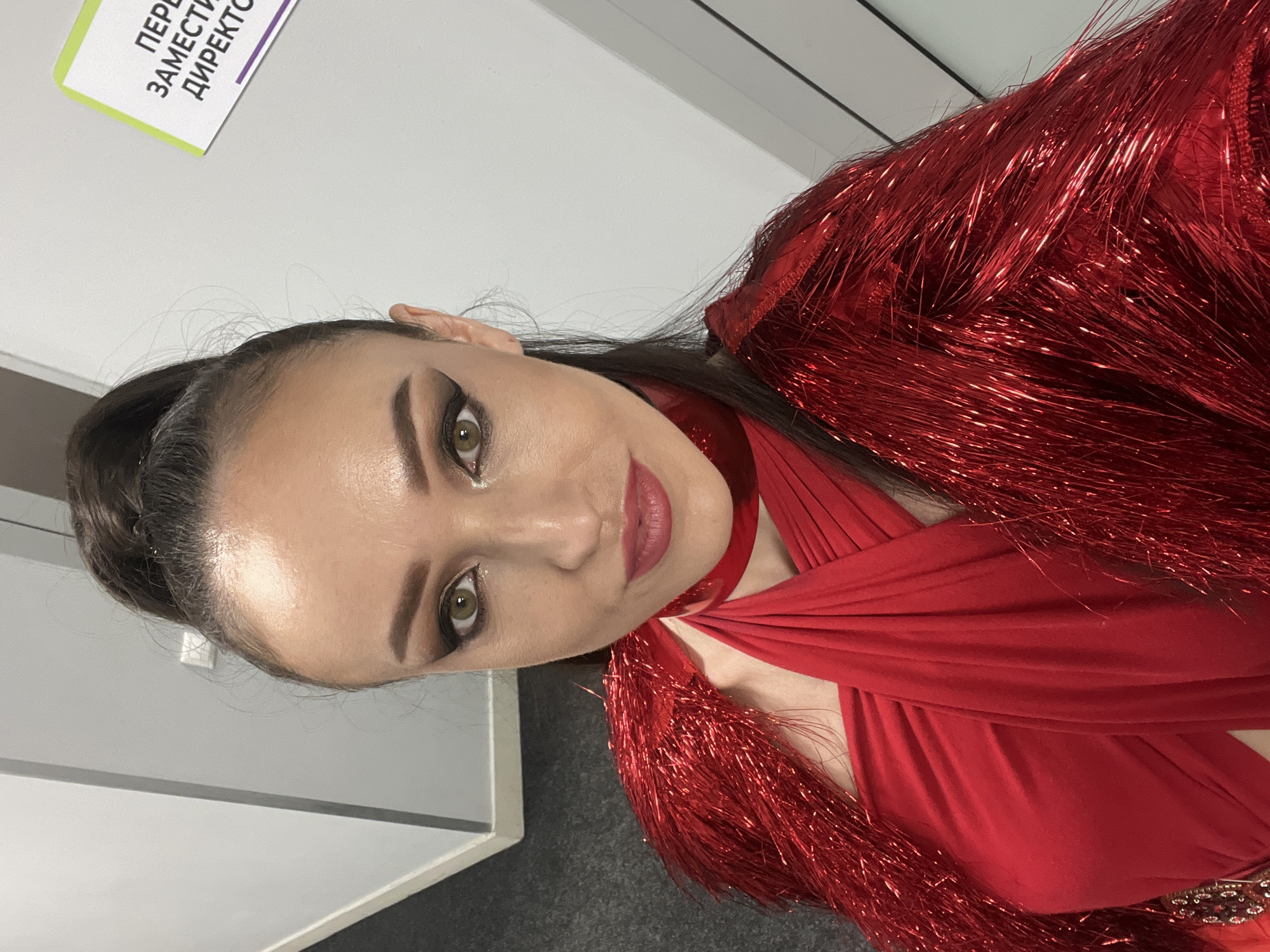 A woman with dark hair pulled back in a high ponytail is centered in the frame, wearing a vibrant red dress with fringed embellishments. She is looking at the camera with a slight smile. The background is dominated by plain white walls, with a sign partially visible in the upper left corner, featuring Russian text in purple and green. The floor appears to be dark gray or black. The photograph was taken indoors, likely a dressing room or backstage area, in Kazan, Russia.