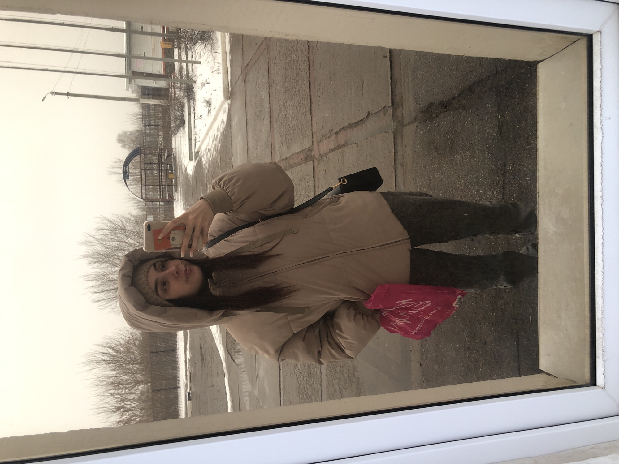 A young woman in a winter hat and hooded beige puffer jacket, with a black crossbody bag, stands inside a building near a white-framed window, taking a selfie with her phone. She holds a pink plastic bag in her left hand. The reflection in the window shows a winter street scene. Bare trees, some snow, and utility poles are visible across the street, in Chaikovsky, Russia. A metal arched structure, possibly part of a park or playground, is also visible in the background.