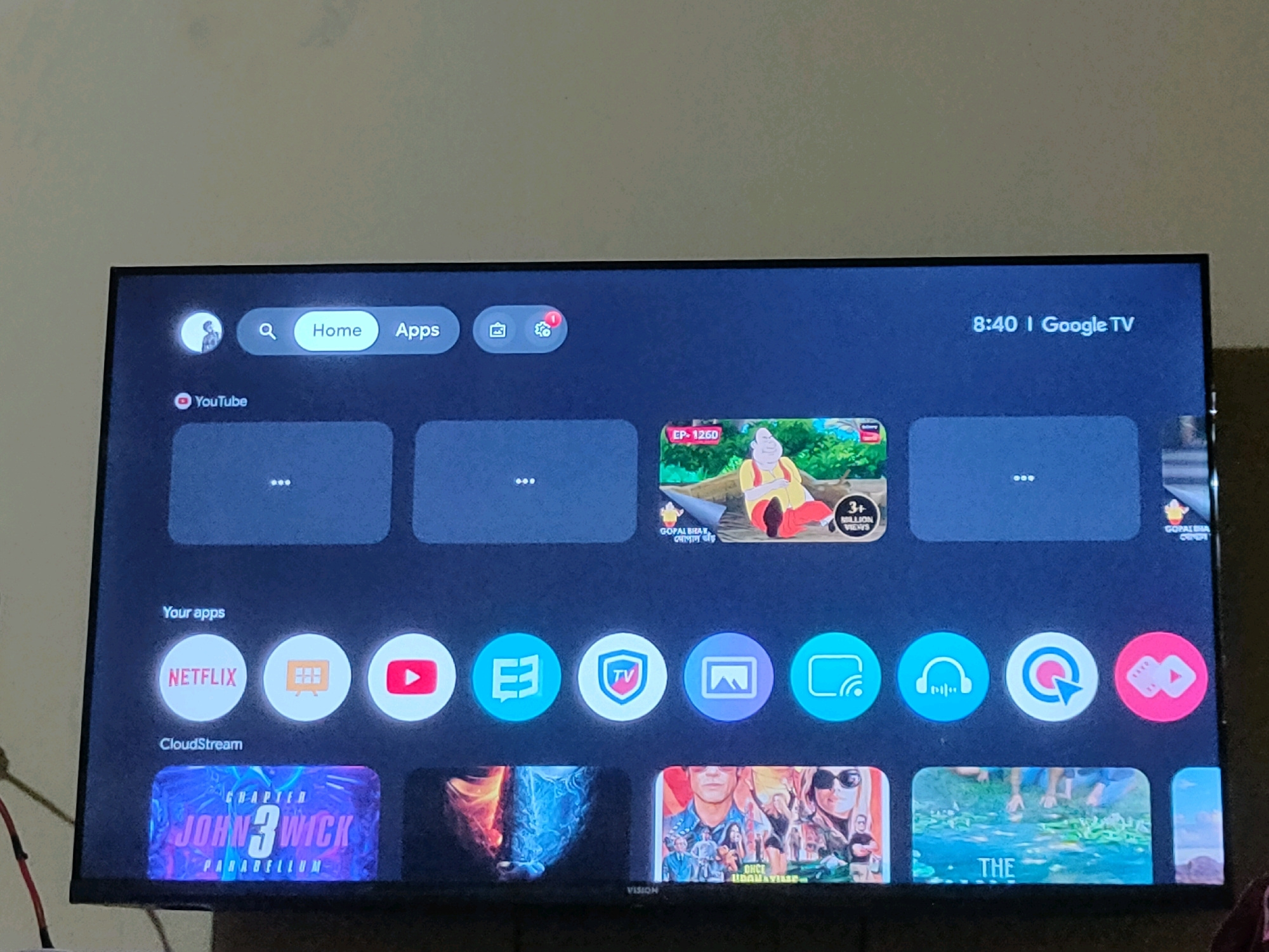 A television screen displays the Google TV interface, showing a mix of apps and content. The interface features a search bar, a "Home" button, and an "Apps" button. Below these, there are rectangular content cards and a row of circular app icons, including Netflix, YouTube, and others. The time "8:40" and "Google TV" are visible in the upper right corner. The television is positioned in a room with a plain wall in the background. The image is taken from a slightly angled perspective. There is no indication of the city of Mohichail, Bangladesh, or any specific environmental context beyond the interior of a room.