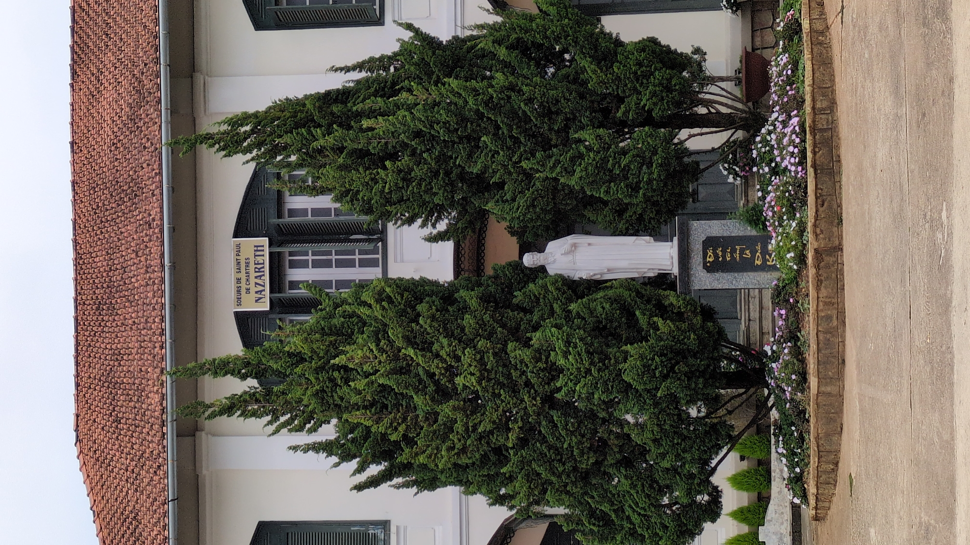 The image shows a statue of a robed figure set among trees and flowers in front of a building. The building is a light color, with green window shutters and a red tile roof. A sign reads "Soeurs de Saint Paul de Chartres Nazareth". The statue appears to be of a religious figure. There is a grey stone behind the statue with writing on it in what appears to be Vietnamese script. The photo may be in the city of Da Lat, Vietnam, given the sign and script on the stone.