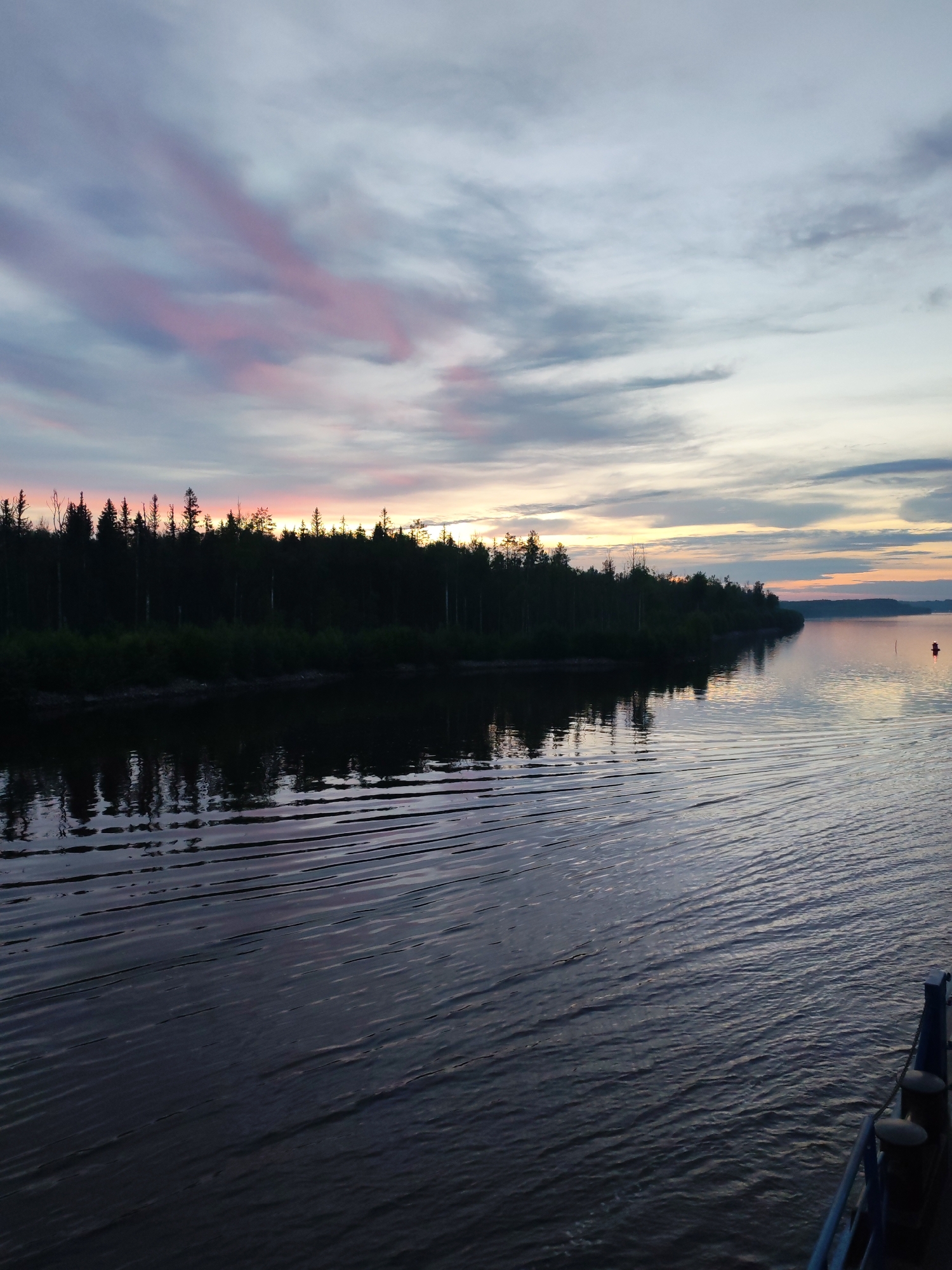 The image depicts a sunset over a wide body of water, likely a river or lake, adjacent to a dense forest. The sky is a gradient of colors, transitioning from pale blue at the top to hues of pink, orange, and yellow near the horizon where the sun is setting. Wispy clouds are scattered across the sky, some tinged with pink. The dark silhouette of a forest lines the distant shore, with the trees appearing tall and dense. The water's surface reflects the colors of the sky and the dark treeline, with visible ripples creating a textured pattern. In the foreground, the dark water fills the lower portion of the image. On the right side of the frame, the edge of a blue boat is visible, with two dark bollards on its deck. A small red buoy is visible in the water in the middle distance. The scene appears serene and natural. While the image is identified as being in Kurdiug, Russia, there are no specific landmarks or identifying features within the image that pinpoint this exact location within the city.
