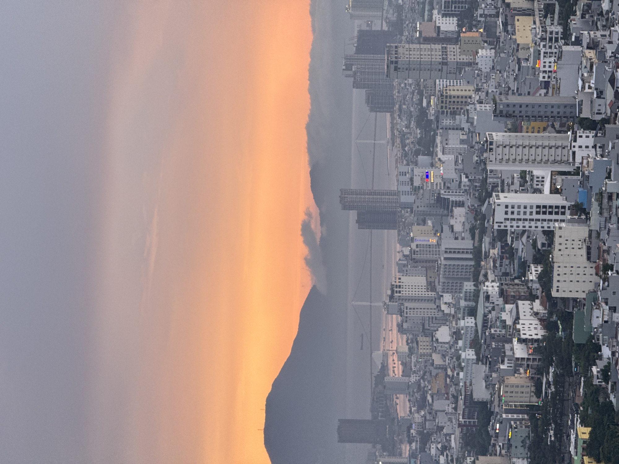 This is an aerial view of the city of Da Nang, Vietnam, at dusk. The sky is a gradient of soft pink and orange from the sunset, transitioning to a pale blue at the top. In the background, a large mountain range is visible, partially obscured by a layer of soft, grey clouds. A suspension bridge spans a body of water in the middle ground, connecting two parts of the city. The foreground is dominated by a dense urban landscape of numerous high-rise buildings, ranging in height and architectural style, with a mix of modern skyscrapers and older structures. Streets and smaller buildings are visible between the larger towers, and scattered patches of dark green trees are interspersed throughout the cityscape. There are no visible people or discernible actions. The overall impression is a sprawling coastal city bathed in the warm, fading light of sunset.