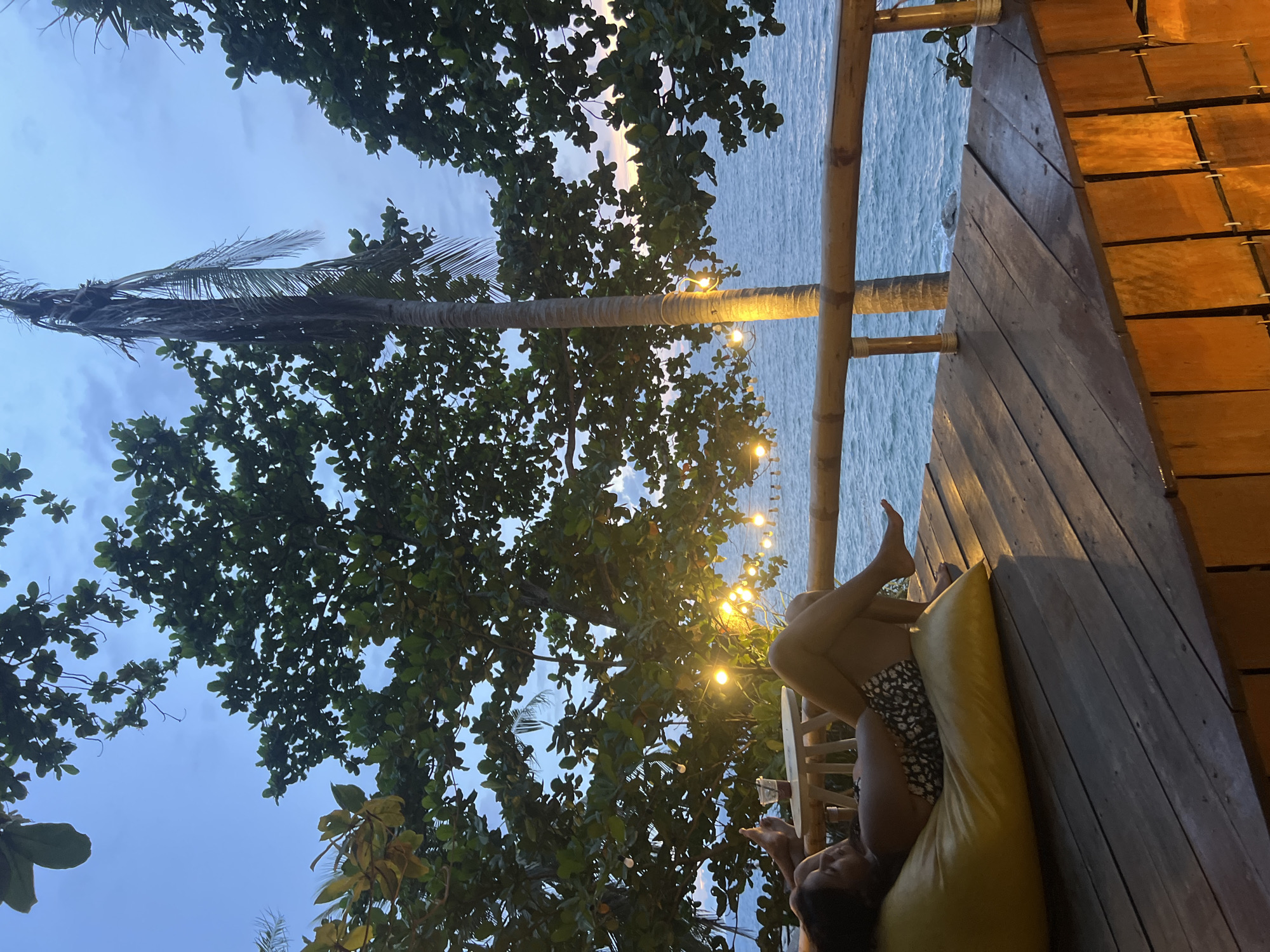 In Karon, Thailand, a person rests on a yellow beanbag chair on a wooden deck overlooking the ocean. The sky is twilight blue with hints of sunset colors near the horizon. Lush green trees with scattered string lights frame the scene, with a palm tree visible in the background. The ocean waves are gentle, and the wooden railing of the deck separates the sitting area from the water. A small table with a drink sits beside the beanbag. The lighting suggests it is evening.