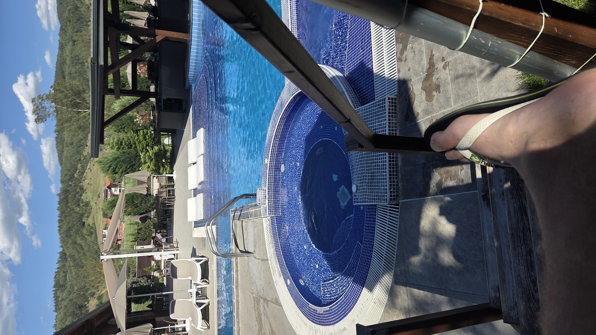 The image depicts a person wearing Havaianas flip-flops standing near a pool and jacuzzi. The setting is outdoors, with a clear blue sky and a mountainous landscape in the background. The pool and jacuzzi are tiled in blue and white mosaics, and lounge chairs are visible. The location appears to be a resort or hotel, situated in a scenic area.