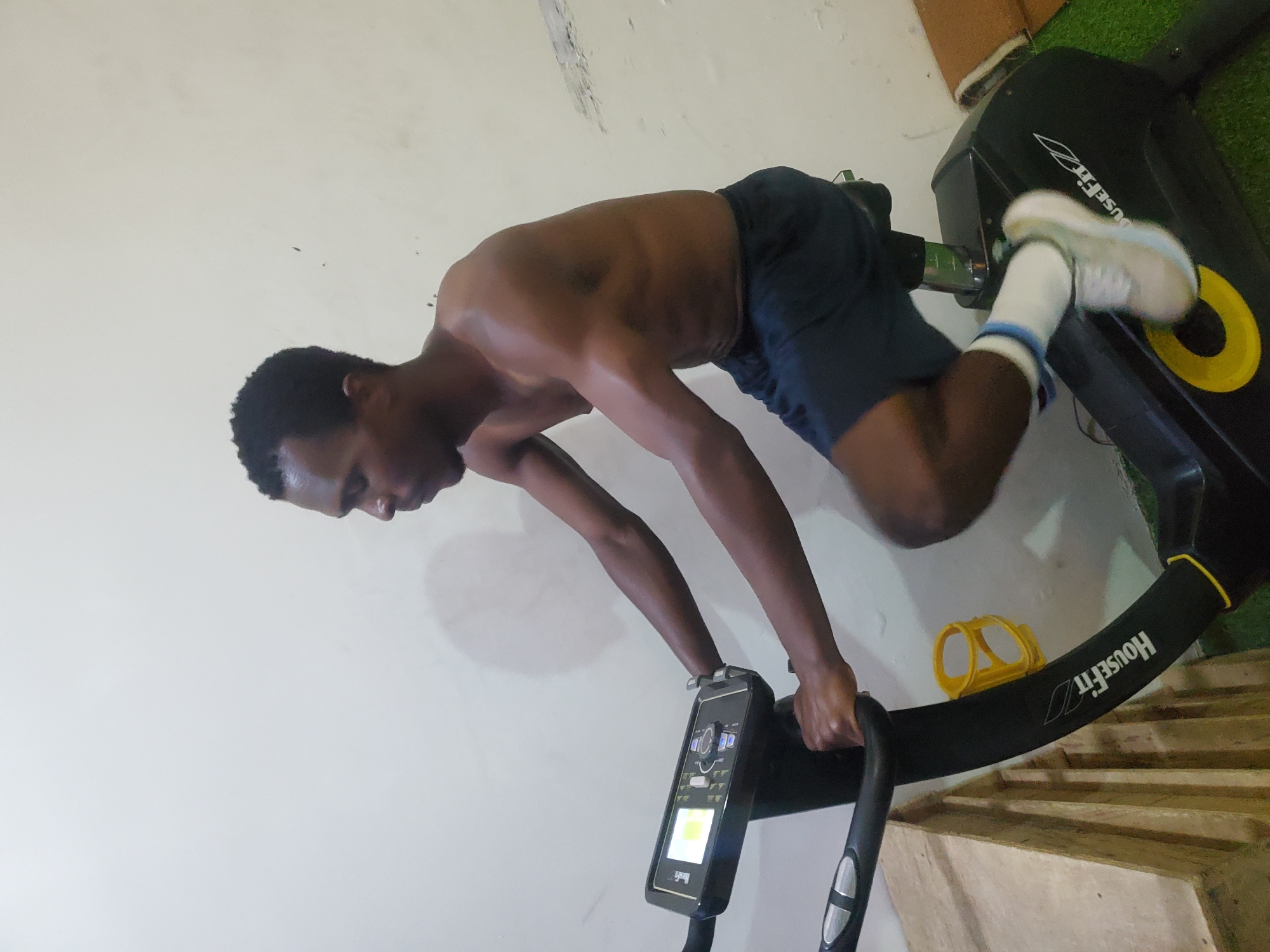 The media file shows a young Black man exercising on a black HouseFit elliptical machine. He is shirtless, wearing dark blue athletic shorts and white socks with blue stripes. His body is angled downwards as he engages in the workout, with his arms supporting his upper body on the handlebars and his legs in motion on the pedals. The elliptical machine has a digital display console. The scene is indoors, with a plain, light-colored wall behind the man and the elliptical. To the right of the elliptical, a section of green artificial turf is visible, and wooden steps or risers are present. A yellow accessory, possibly a water bottle holder, is attached to the elliptical frame. The lighting suggests it is daytime, and the overall mood is one of focused physical exertion. The visible text on the elliptical reads "HouseFit".