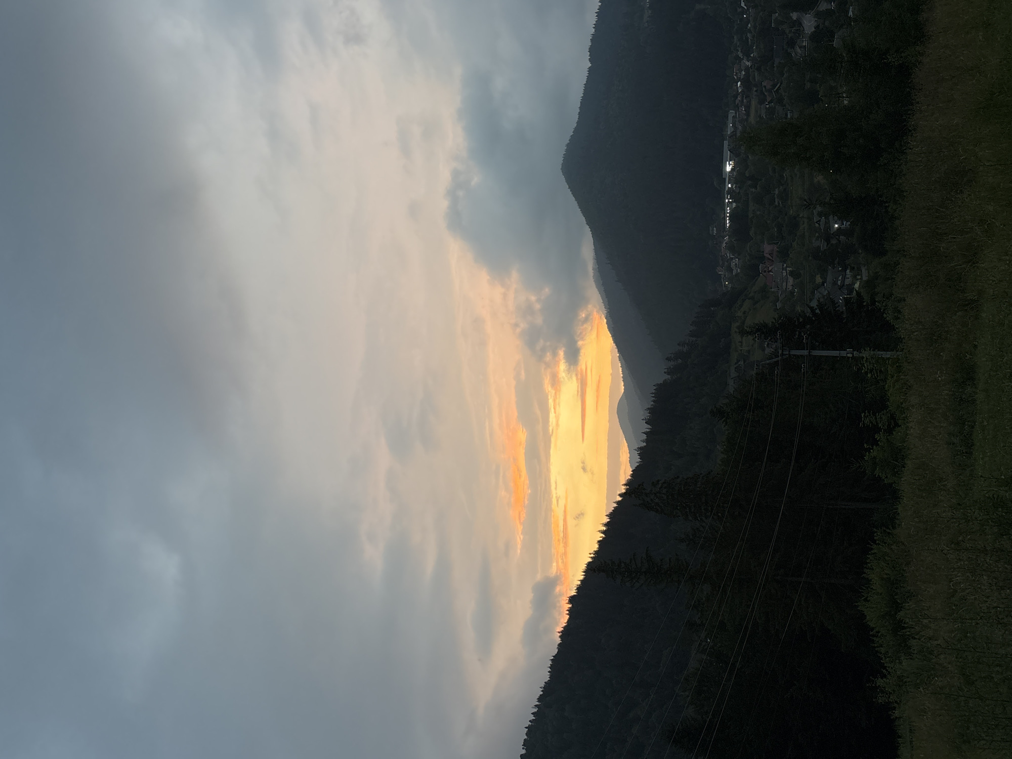 The media file captures a dramatic sunset over a mountainous landscape in Tatariv, Ukraine. The scene is dominated by a vast sky filled with both dark, heavy clouds and patches of vibrant orange and yellow hues, indicating the sun's descent. Silhouetted against this colorful sky are the dark, tree-covered slopes of mountains, one of which extends diagonally across the frame, obscuring much of the view.
Below the mountains, a small village or town is visible, with scattered lights suggesting the early evening hours. The architecture is not clearly discernible due to the distance and low light conditions. Power lines stretch across the middle ground, adding a man-made element to the natural scenery. The foreground features more dark foliage, possibly trees or tall grass, further emphasizing the natural setting.
The time of day is clearly sunset, with the dramatic lighting and cloud formations being key indicators. The weather appears to be mixed, with storm clouds present alongside the sunlit areas, creating a moody and visually striking atmosphere. The emotion conveyed is one of awe and perhaps a hint of foreboding due to the dark clouds. There is no visible text in the image.
