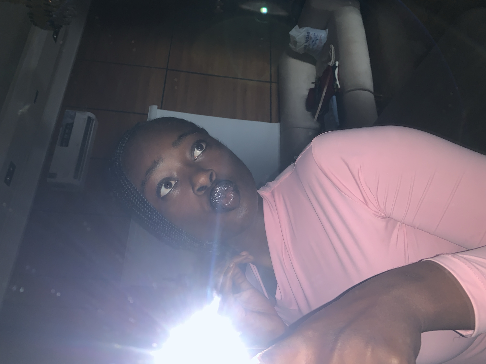 A young Black woman with dark braided hair and glittery lipstick is captured in a close-up, off-center, selfie-style shot. She is wearing a pink, off-the-shoulder long-sleeved top. The bright light source, likely a flash, is positioned to her left, creating a strong glare and lens flare across the left side of the image, obscuring some details. Her head is tilted, and her eyes are wide open, looking slightly off-camera to her right. Her left hand is raised to her face, near her mouth.
The background appears to be an indoor setting, possibly a living room or common area. Behind her, a light-colored wall or screen is visible, and to the right, a dark wooden paneled wall is present. Mounted on the wall is a white air conditioning unit. Further to the right, a sofa with beige upholstery is partially visible. On the sofa, there are several items, including what appear to be shoes or bags. A white plastic bag with some writing on it sits on the sofa. The overall lighting is dim, with the exception of the direct light source. The location is described as Gura Top, Nigeria.