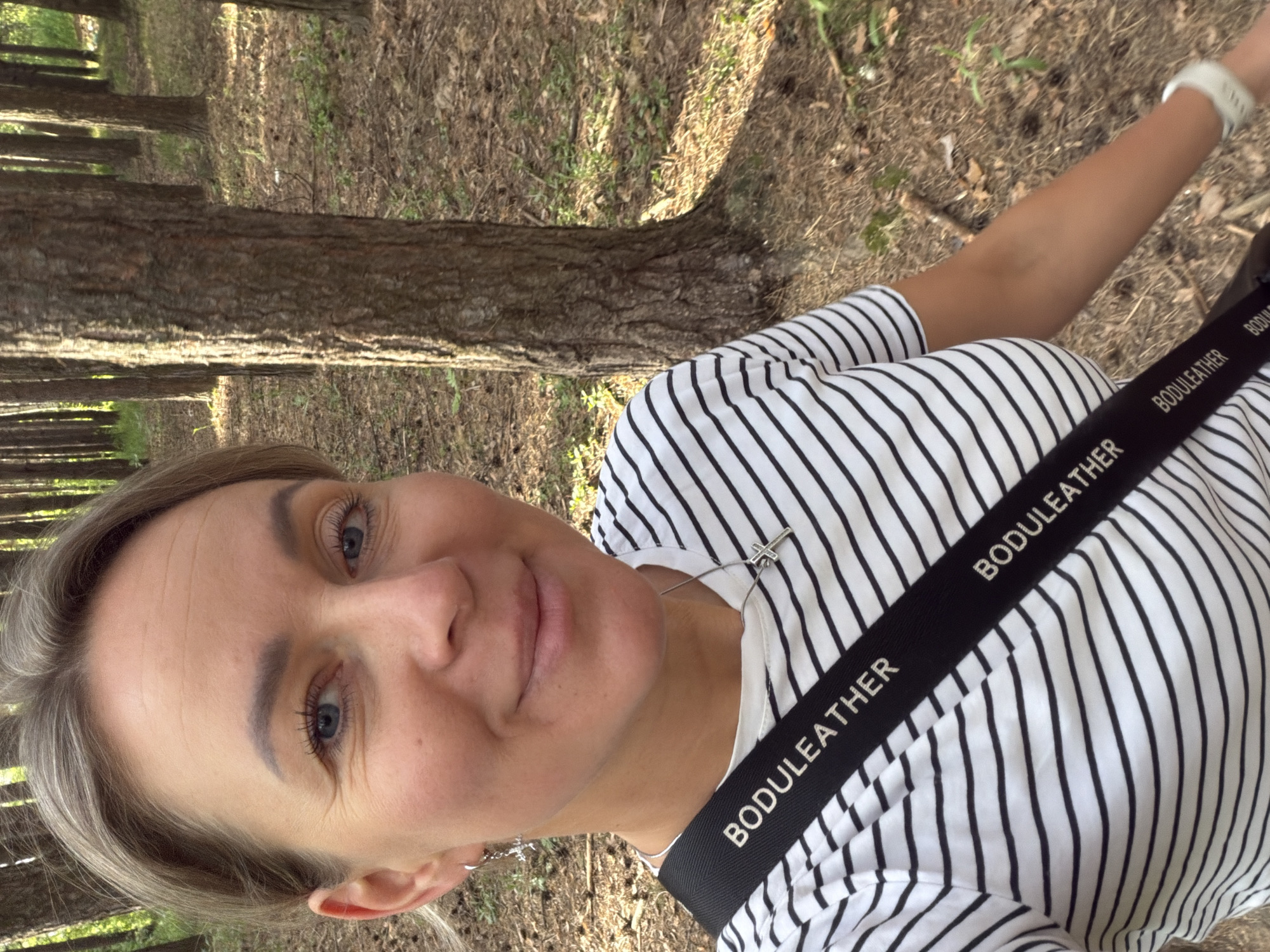 A fair-skinned woman is facing the camera in an outdoor setting. She has blue eyes and blonde hair. She is wearing a white and black striped top and a black strap with "BODULEATHER" printed on it. She also wears a white wristband. The background shows tree trunks and ground covered with grass and leaves. It is likely a forested area. While the image does not explicitly show Chaikovsky, Russia, the user's prompt suggests it is the location.