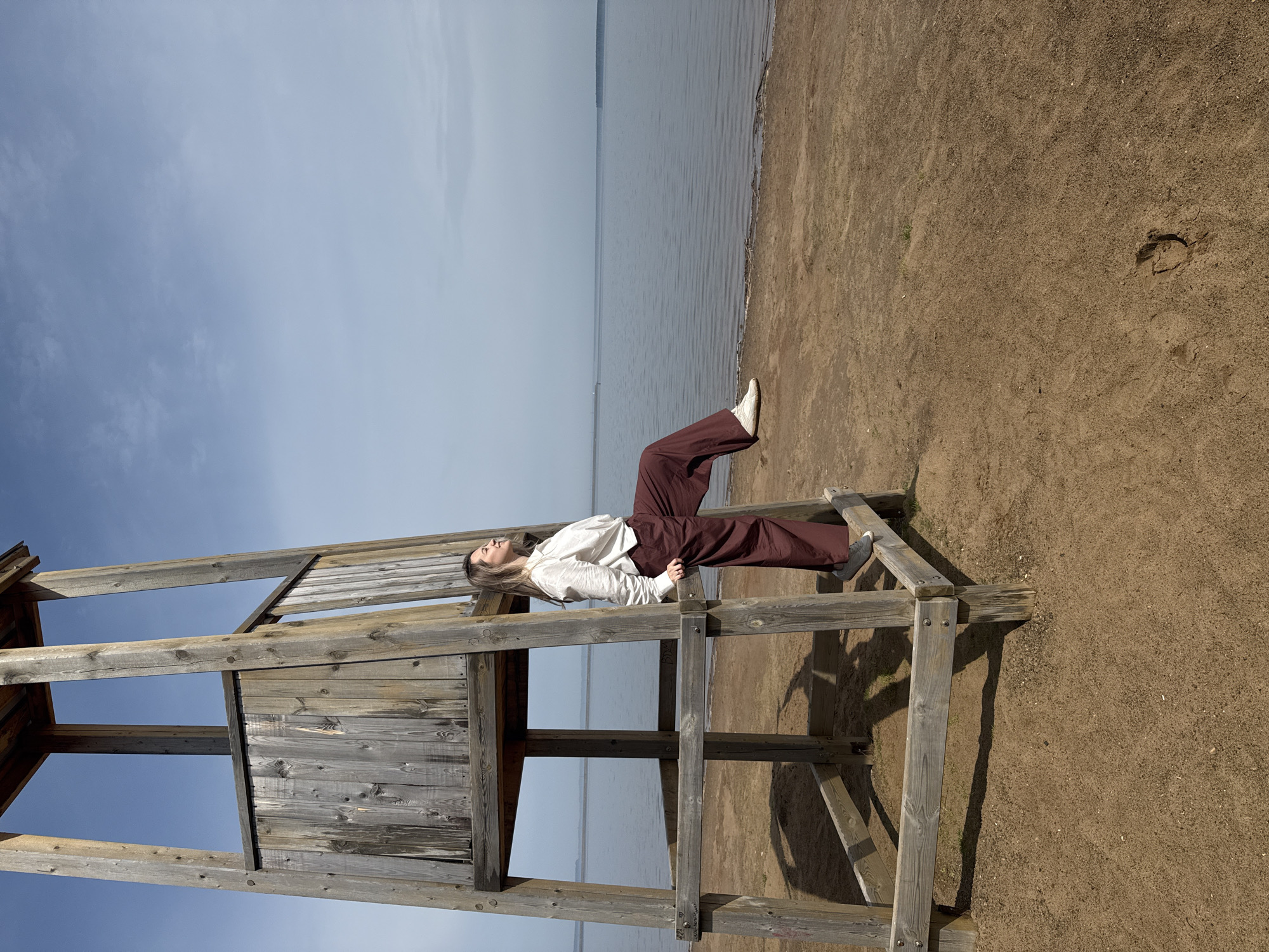 A person lies on a wooden structure on a sandy beach, facing towards a body of water under a partly cloudy sky. The person is wearing brown pants and a light-colored shirt, with white sneakers. The wooden structure appears to be part of a lifeguard stand. The beach is sandy with some visible footprints. The water is calm, and the sky is a mix of blue and light gray clouds. The location is Chaikovsky, Russia.