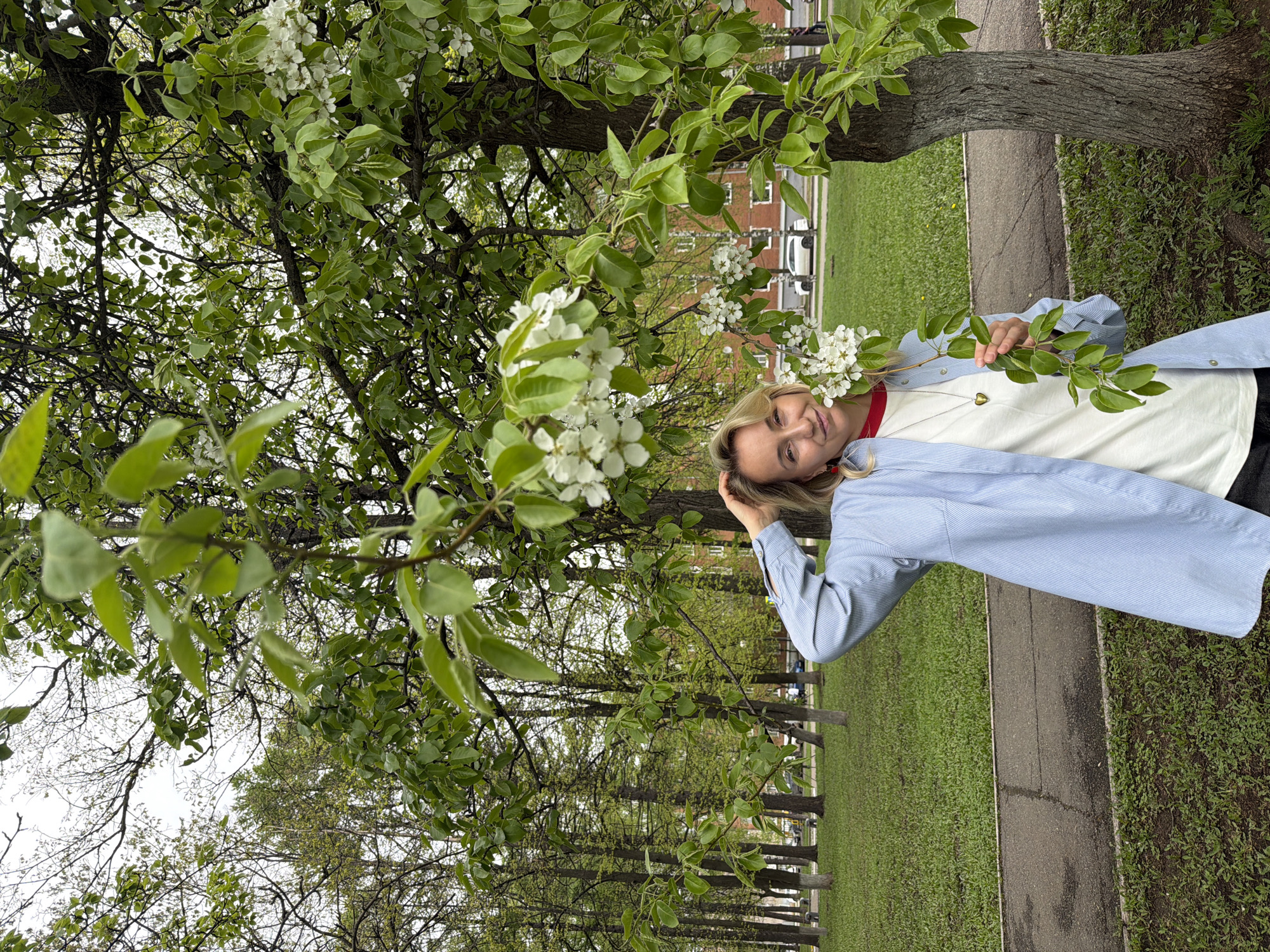 A woman stands beside a tree in a grassy area next to a paved walkway. She is fair-skinned with blonde hair and wears a light blue, striped shirt over a white t-shirt. She is touching her head with one hand and is near flowering branches. Buildings and cars are visible in the background. The location is Izhevsk, Russia.