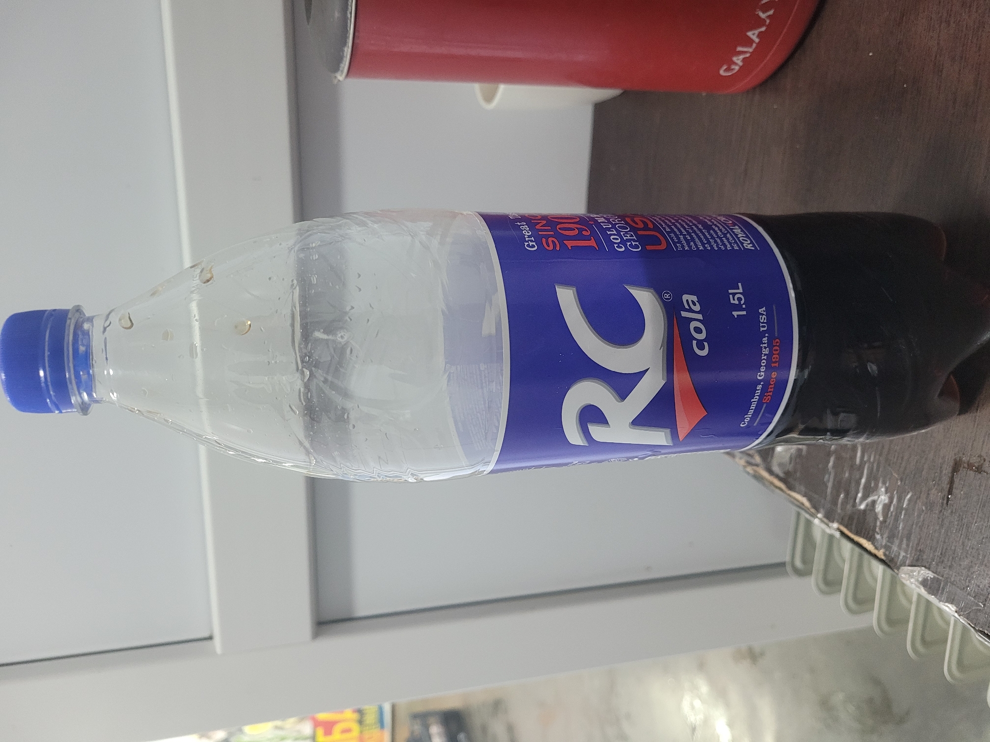 In Mytishchi, Russia, a clear plastic bottle of RC Cola, labeled "cola 1.5L" and "Since 1905", stands upright on a dark wooden surface. The bottle is partially filled with a dark brown liquid. A blue cap secures the top of the bottle. To the right of the bottle, a red cylindrical object with the word "GALAXY" in white lettering is visible. The background consists of a light-colored, textured wall and a hint of a white railing.