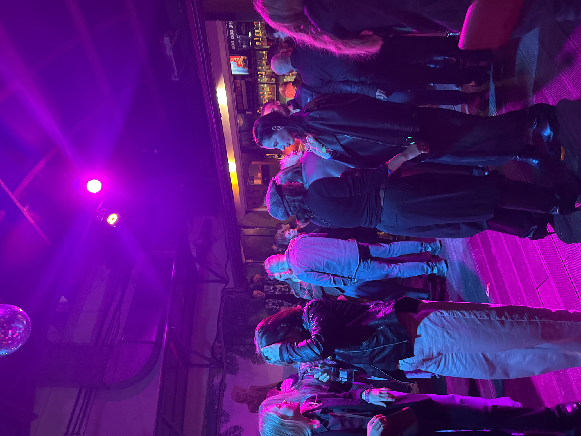 The image depicts a dimly lit nightclub or bar, bathed in vibrant purple and pink lighting, likely from a disco ball and stage lights. A diverse group of people are gathered, some standing and talking, others appearing to be dancing or socializing. The environment suggests a lively, urban nightlife setting. The clothing of the individuals ranges from casual to slightly more formal attire, with some individuals wearing leather jackets. In the background, a bar area is visible with shelves stocked with bottles, and a screen displaying text in Russian, possibly advertising or promotions. The floor appears to be made of wooden planks. The overall atmosphere is one of a social gathering or event.