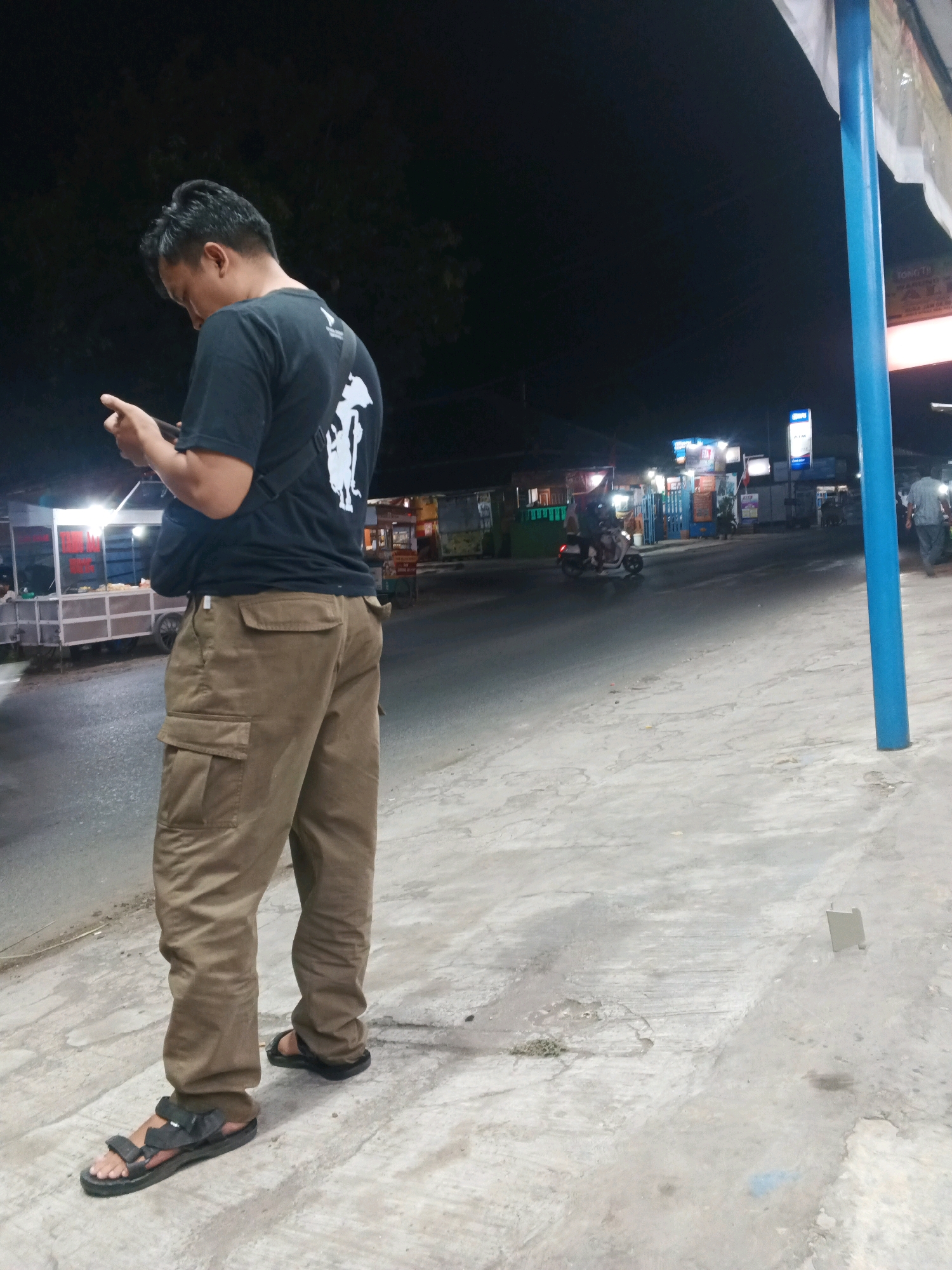 A man stands on a sidewalk at night, looking at his phone. He is wearing a black t-shirt and tan cargo pants with black sandals. To his left, a street vendor cart is visible with lights illuminating the food displayed. Across the street, there are several businesses with illuminated signs, including what appears to be an ATM. A scooter is seen driving on the wet road. The overall environment suggests a street scene in Taman, Indonesia, likely a commercial or residential area with active street life even at night. A blue pole stands on the right side of the frame, partially obscuring a white tarp.