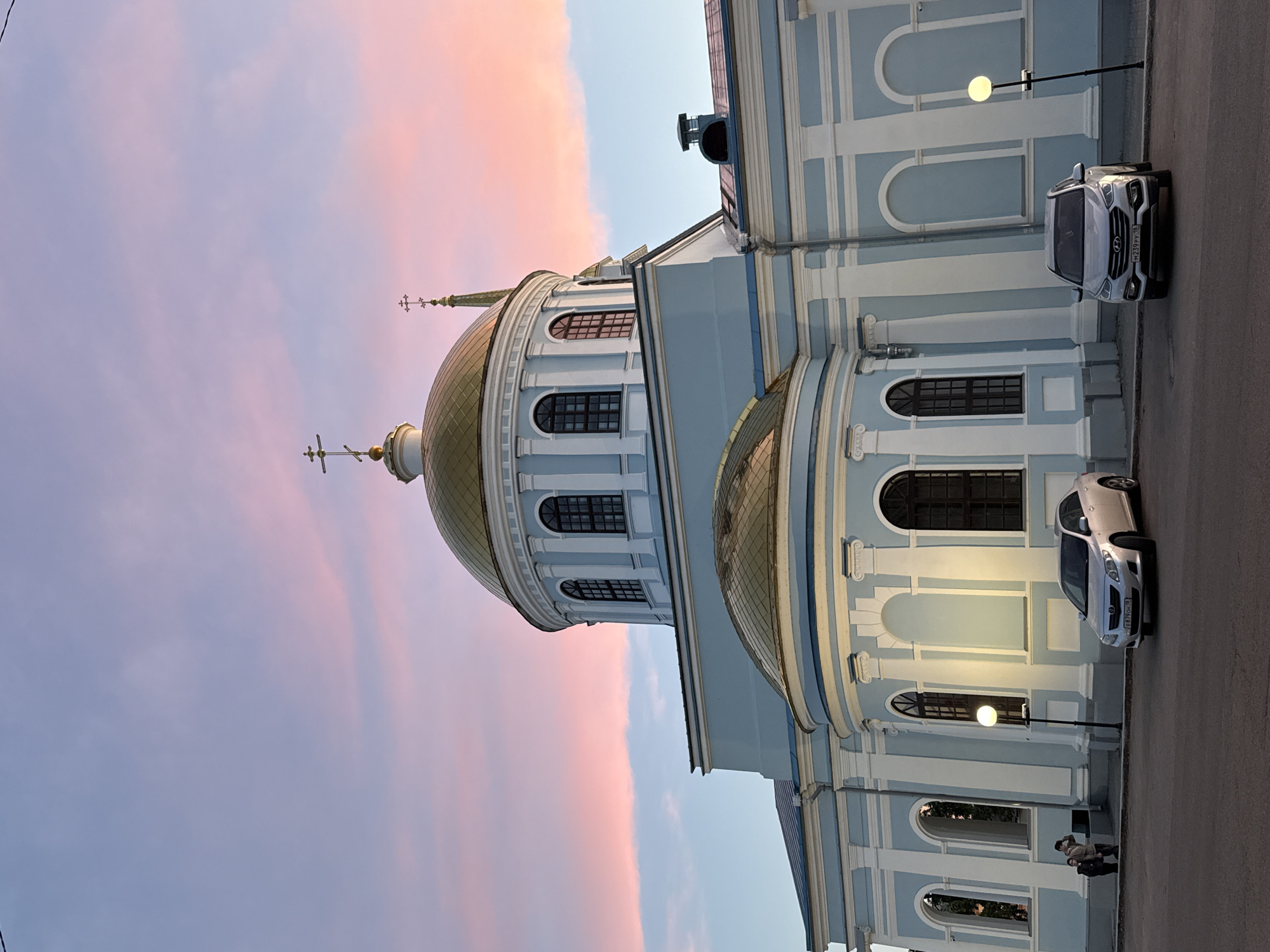 The image captures a serene evening scene in Votkinsk, Russia, featuring a grand Orthodox church or cathedral under a sunset sky. The magnificent building is characterized by its light blue facade accented with prominent white architectural details, including columns, arched window frames, and cornices. Several golden, onion-shaped domes crown the structure, each topped with an ornate cross. The largest central dome features multiple arched windows.
The sky provides a striking backdrop, displaying a broad band of soft pink and orange clouds against a lighter blue, typical of late afternoon or early evening. The ambient light suggests either sunset or sunrise, with streetlights already illuminated, casting a warm, circular glow.
At ground level, a paved street or parking area runs alongside the church. Two light-colored cars are parked there; one is a silver SUV (likely a Hyundai Santa Fe) with a visible Russian license plate reading "H239PY". Two individuals are also visible near the base of the church, standing or casually walking beneath one of the glowing streetlights. The overall impression is one of peaceful beauty, with the historic architecture bathed in the warm hues of the fading day.