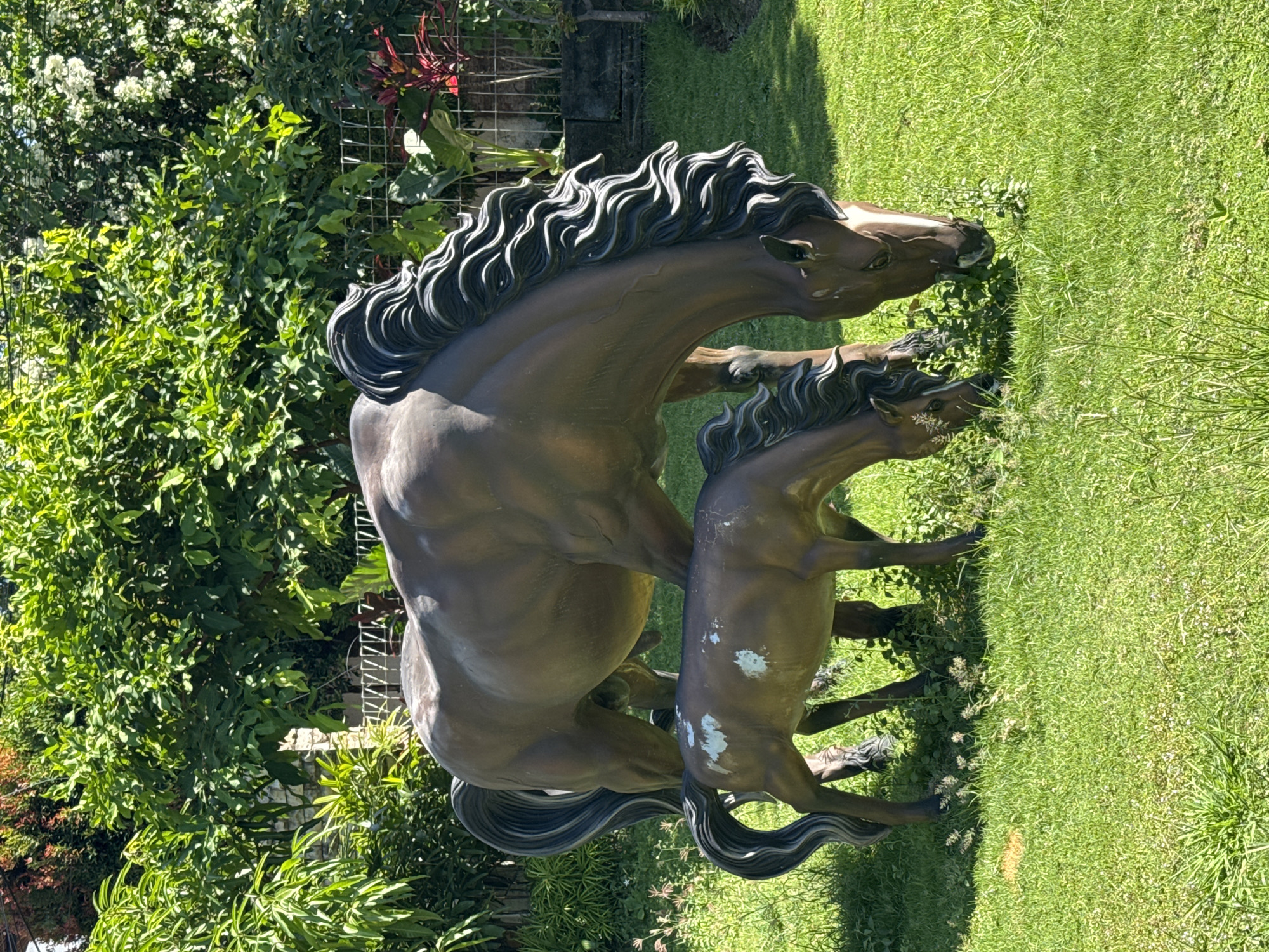A statue of a mare and foal stands on a patch of grass in North Kuta, Indonesia. The statue is bronze colored. The mare is larger, and her head is bent towards the ground as if grazing. The foal is smaller and stands beside the mare. The background consists of green foliage and a small section of what appears to be a fence.