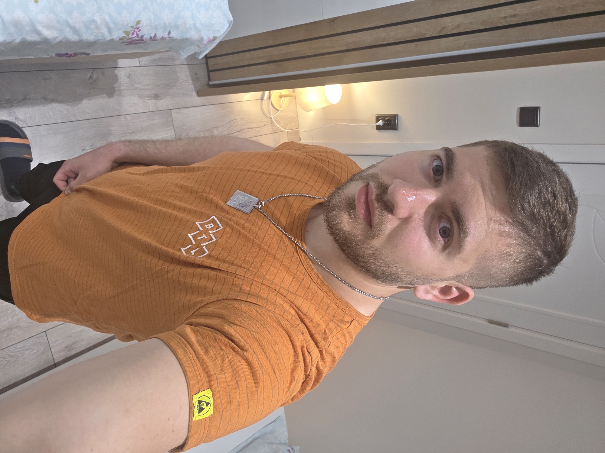 A man is pictured from a low angle. He wears an orange T-shirt with a logo on the chest and a yellow sticker on the sleeve. He has a chain necklace with a pendant. He has short brown hair and a short beard. He is indoors, possibly in a bedroom. A bed with floral sheets is visible in the background. The location is impossible to determine.
