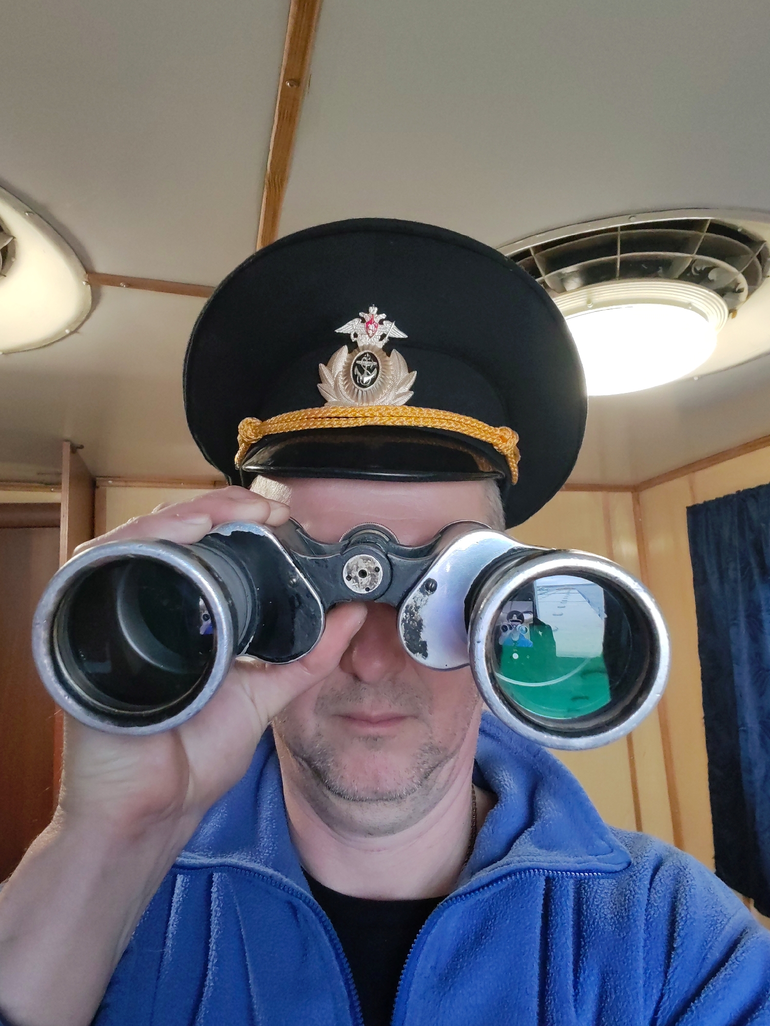 A man, seen from the chest up, is shown looking directly forward through a pair of binoculars. He appears to be of middle age with visible stubble on his chin and cheeks. He is wearing a black uniform cap adorned with a gold braided band and a silver, red, and gold emblem featuring a double-headed eagle and an anchor, characteristic of Russian naval or maritime insignia. He wears a blue fleece jacket over a dark shirt.
The man holds a pair of worn silver and black binoculars to his eyes, his hands cupping the barrels. In the reflection on the right objective lens of the binoculars, a bright, distant outdoor scene, possibly a ship's deck or horizon, is faintly visible, along with a distinct green surface.
The setting appears to be an interior space, likely a cabin or bridge of a vessel, suggested by the maritime uniform and binoculars. The ceiling is light-colored, with wooden beams running across it. Two circular light fixtures are visible, one of which has a prominent gridded vent around it. On the right side of the frame, light wooden paneling and a dark blue curtain can be seen. The overall impression is that of daytime due to the interior lighting. No visible text is present in the image. The location context is Bol'shie Mkhi, Russia.