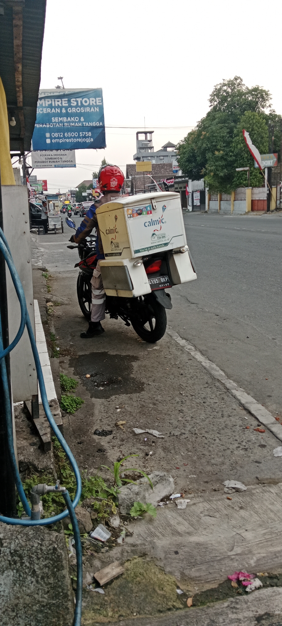 A person wearing a red helmet and a brown and blue jacket is sitting on a motorcycle. The motorcycle has two insulated boxes attached to the rear. The left box is yellow with the "calmic" logo and "You're in safe hands" text. The right box is white, also with the "calmic" logo and "You're in safe hands" text, and a graphic of a kite. The motorcycle's license plate reads "4195 BKP".
The motorcycle is parked on a sidewalk next to a street. The street appears to be in an urban area with buildings and trees visible in the background. There is a sign in the background that reads "EMPIRE STORE ECERAN & GROSIRAN SEMBAKO & PERABOTAN RUMAH TANGGA" which translates to "EMPIRE STORE RETAIL & WHOLESALE GROCERIES & HOUSEHOLD FURNITURE". The location is identified as Maguwoharjo, Indonesia. The sidewalk is made of concrete and shows signs of wear and tear, with some vegetation growing in cracks and patches of standing water. There is also some debris on the sidewalk, including plastic wrappers and a discarded cigarette butt. A blue hose is visible in the foreground, connected to a water tap. The sky is overcast.