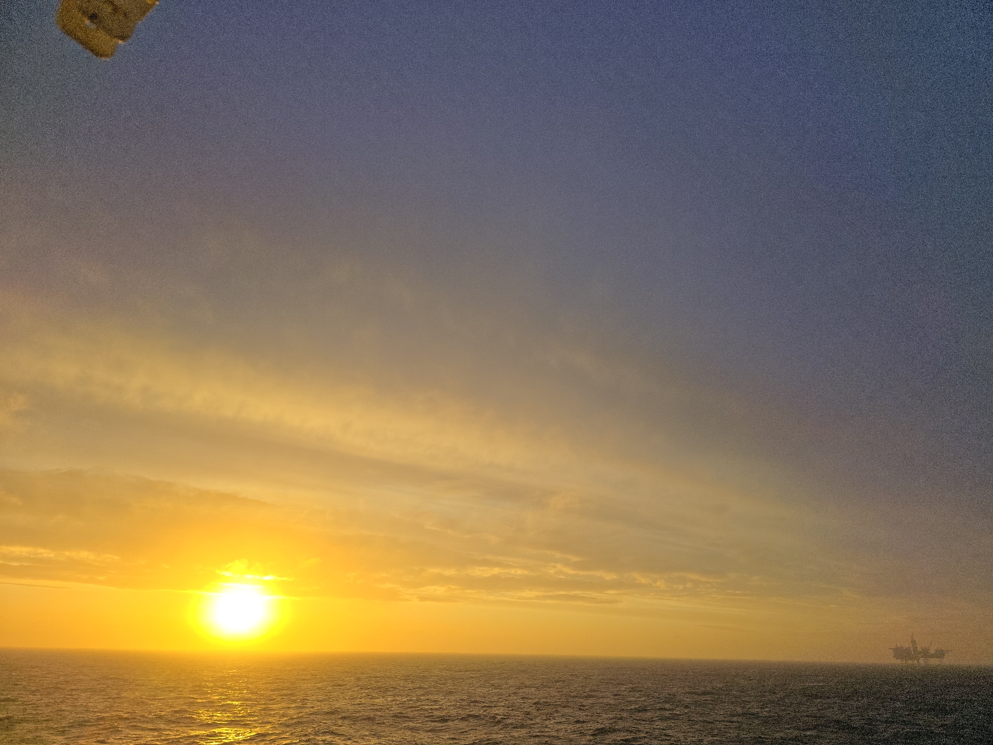 The image captures a sunset over the ocean. The sun is low on the horizon, emitting a bright, golden light that reflects on the water's surface. The sky transitions from a vibrant yellow near the horizon to a deep blue higher up. There are a few wispy clouds scattered across the sky. In the distance, on the right side of the frame, there appears to be an oil rig. The location is Unknown, Unknown.