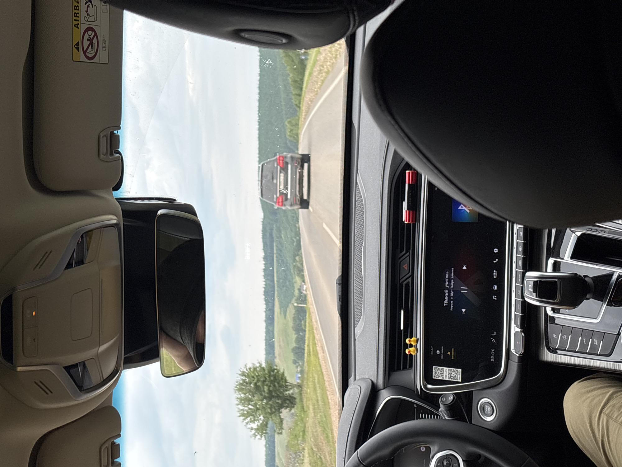 The image shows the interior of a car driving on a road, with a landscape visible through the windshield. In the foreground, part of the dashboard and seats are visible. On the dashboard, there is a digital display screen showing music information and a QR code. Two yellow duck figures are placed on the dashboard near the air vents. The steering wheel is partially visible on the left.
In the middle ground, a dark-colored SUV is driving ahead on the same road. The road itself appears to be asphalt and is flanked by grassy shoulders. The background features rolling green hills covered with trees, indicating a rural or semi-rural environment. The sky is overcast with some patches of blue. The overall lighting suggests daytime.
The image is taken from the perspective of a passenger or driver inside the car. The text on the dashboard display appears to be in Russian, with the song title "Тёмный учитель" (Dark Teacher) and artist "Король и Шут-Театр демона" (Korol i Shut - Demon Theater) visible. The temperature is displayed as 22.0°C.
The location is stated to be Дюсьметьево, Russia. This context suggests the scene is taking place on a road within or near this locality.