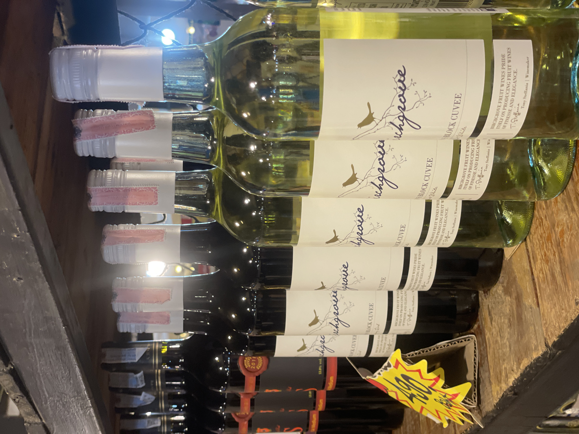 The image displays a close-up view of several bottles of wine arranged on a wooden shelf in what appears to be a retail setting. The bottles are positioned in a row, with some being red wine and others white wine, distinguishable by the color of the liquid visible through the glass. The labels on the white wine bottles are white with a cursive script logo and an illustration of a bird on a branch. The red wine bottles have darker labels. Some of the bottles have metallic-colored caps with pink and white paper seals. To the left of the wine bottles, a bright yellow and red price tag with the number "490" in a stylized font is visible, attached to the shelf. The background shows a wooden shelf or counter and a dimly lit area beyond the bottles. The overall impression is of a wine display in a shop. The presence of the price tag suggests a commercial environment. The lighting in the image is somewhat dim, with reflections visible on the glass bottles. The image does not contain any people.